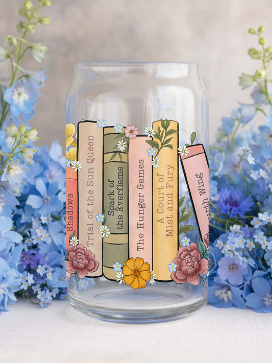 Clear glass tumbler featuring pastel-colored book spines like “A Court of Mist and Fury” and “The Hunger Games,” surrounded by hand-drawn florals and blue blossoms for a dreamy book lover aesthetic.