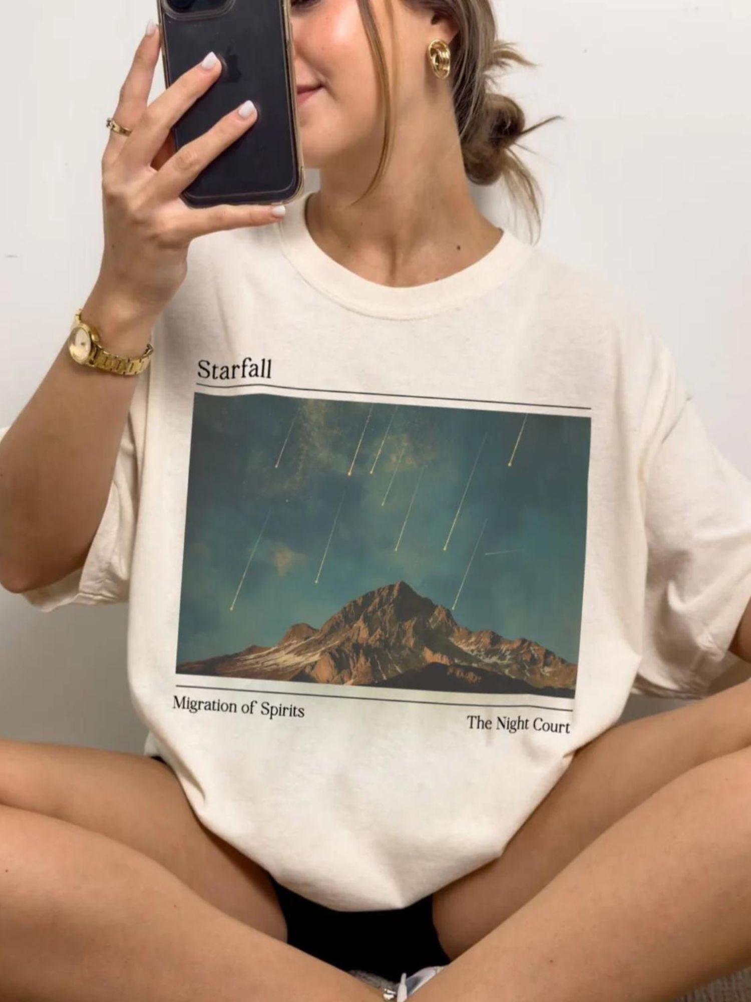 Girl sitting down wearing a natural color tshirt. The graphic design says "Starfall The Night Court" with an image of falling stars over a mountain