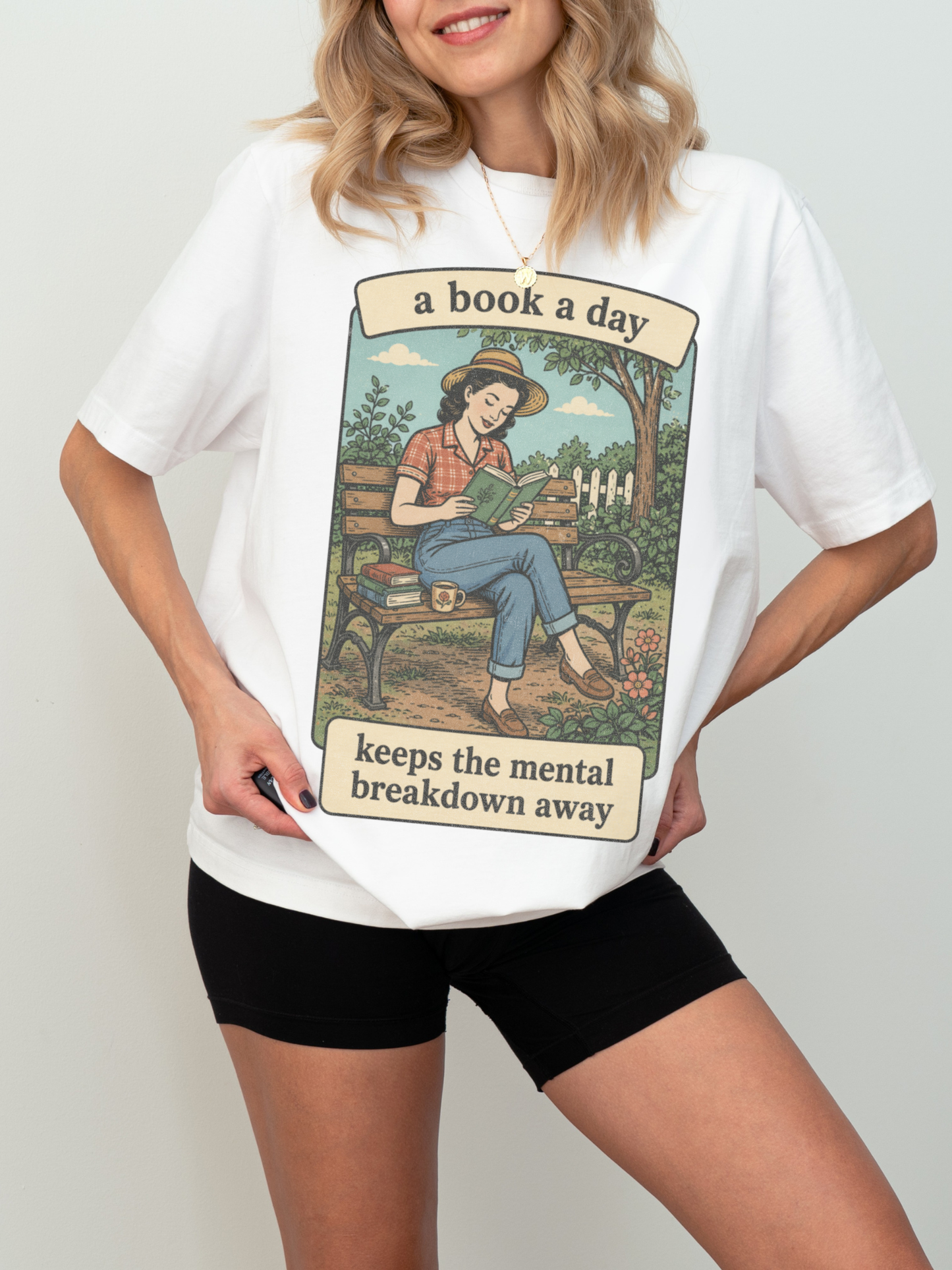 Woman wearing a white oversized t shirt with retro book lover design of woman reading on bench and text a book a day keeps the mental breakdown away