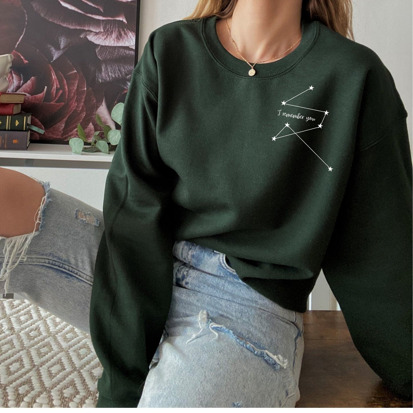 woman wearing a forest green sweatshirt with a white Addie LaRue constellation and the phrase I remember you