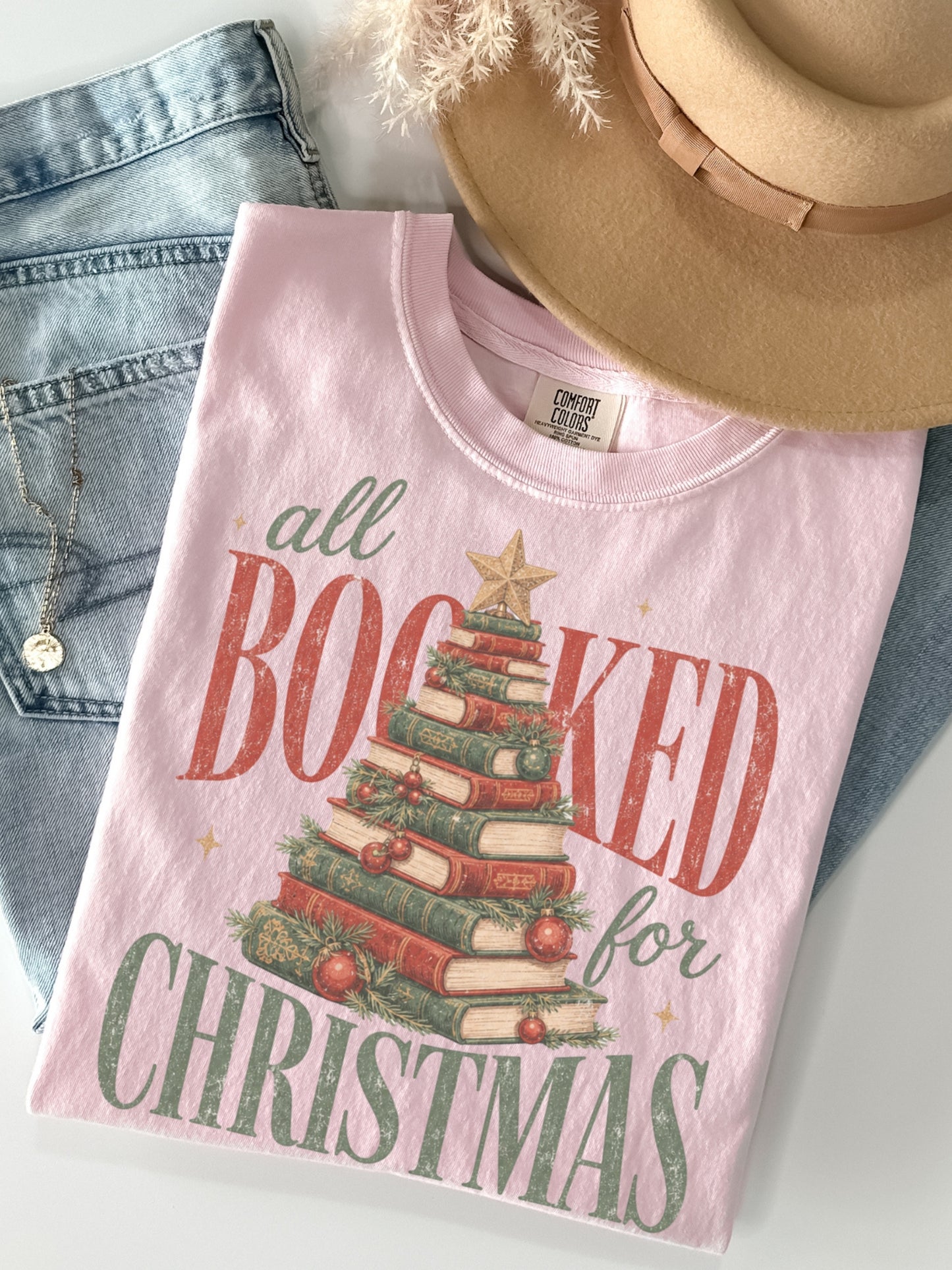 Blossom Comfort Colors shirt featuring an All Booked for Christmas graphic with a Christmas tree made from stacked books, evergreen greenery, red ornaments and gold star, cozy holiday book lover t shirt