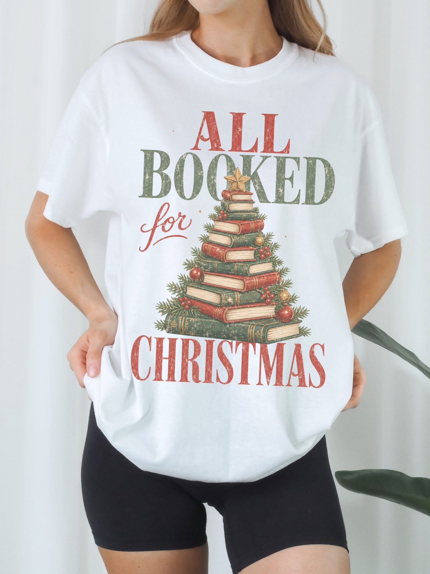 White Comfort Colors shirt modeled with an All Booked for Christmas graphic featuring a festive Christmas tree made from stacked vintage books with evergreen branches, red and gold ornaments, berries and a gold star