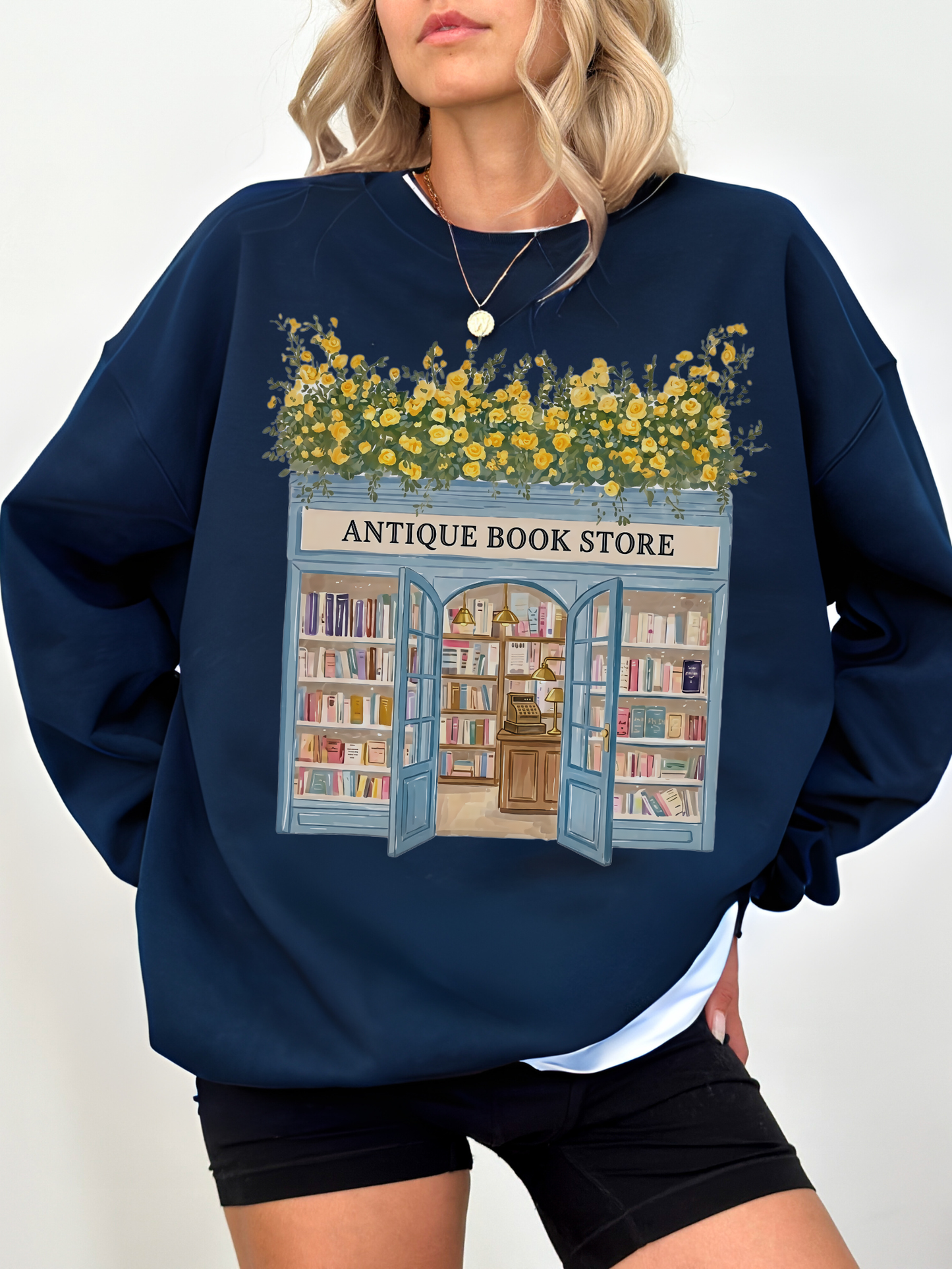Woman wearing a navy blue sweatshirt with a vintage bookstore design featuring open doors, bookshelves, and yellow flowers styled with black shorts in a casual look