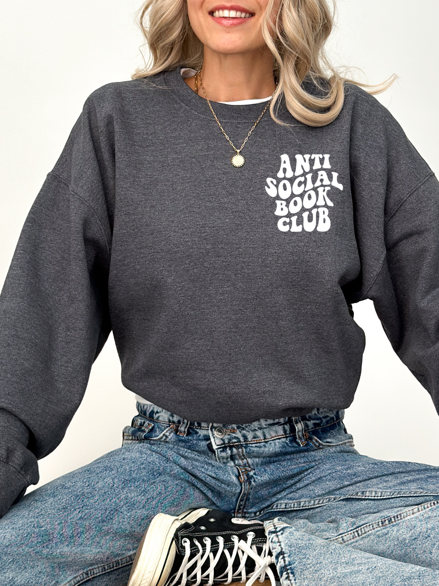 woman sitting cross-legged wearing dark heather sweatshirt with anti social book club text and jeans