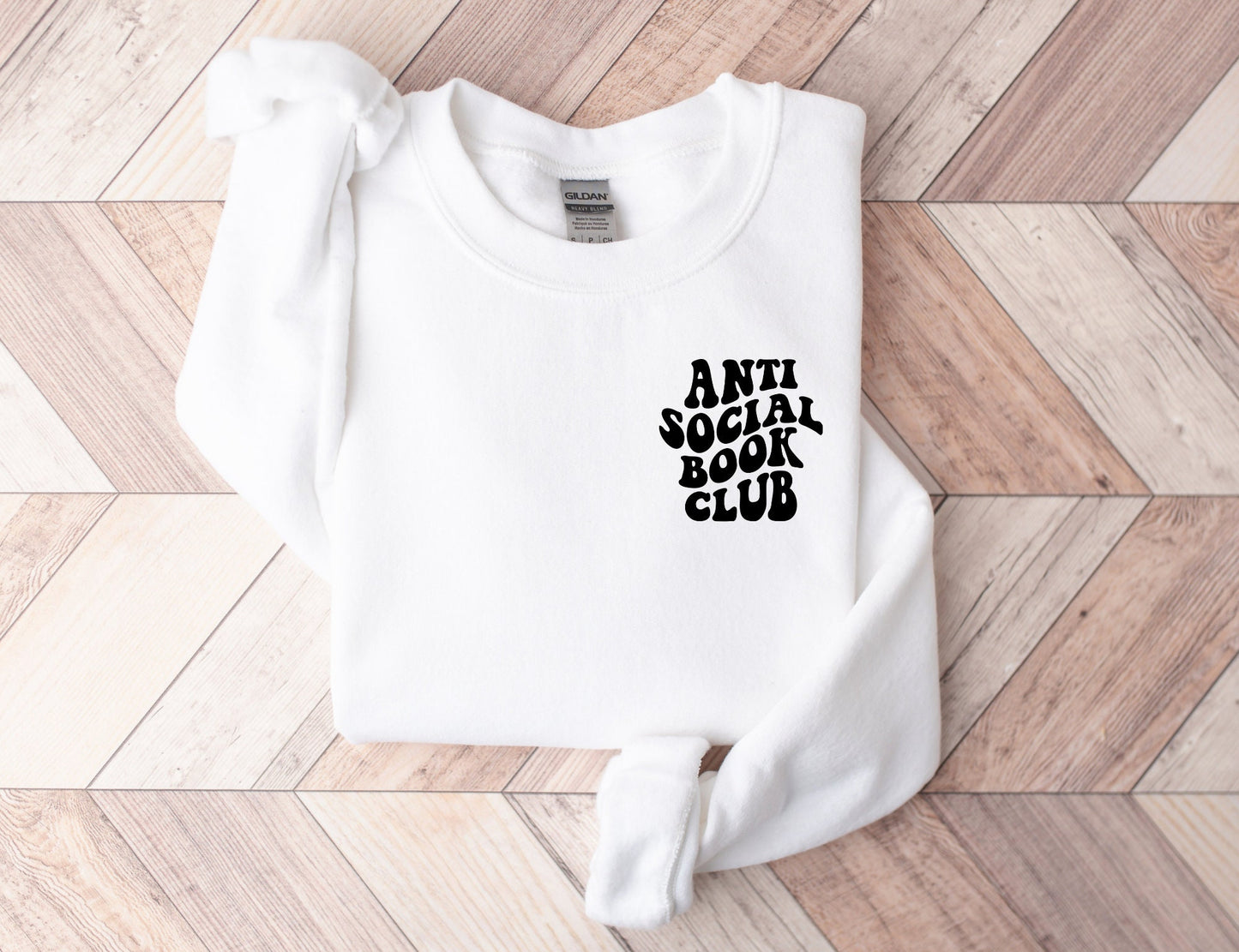folded white sweatshirt with anti social book club text on wood herringbone background