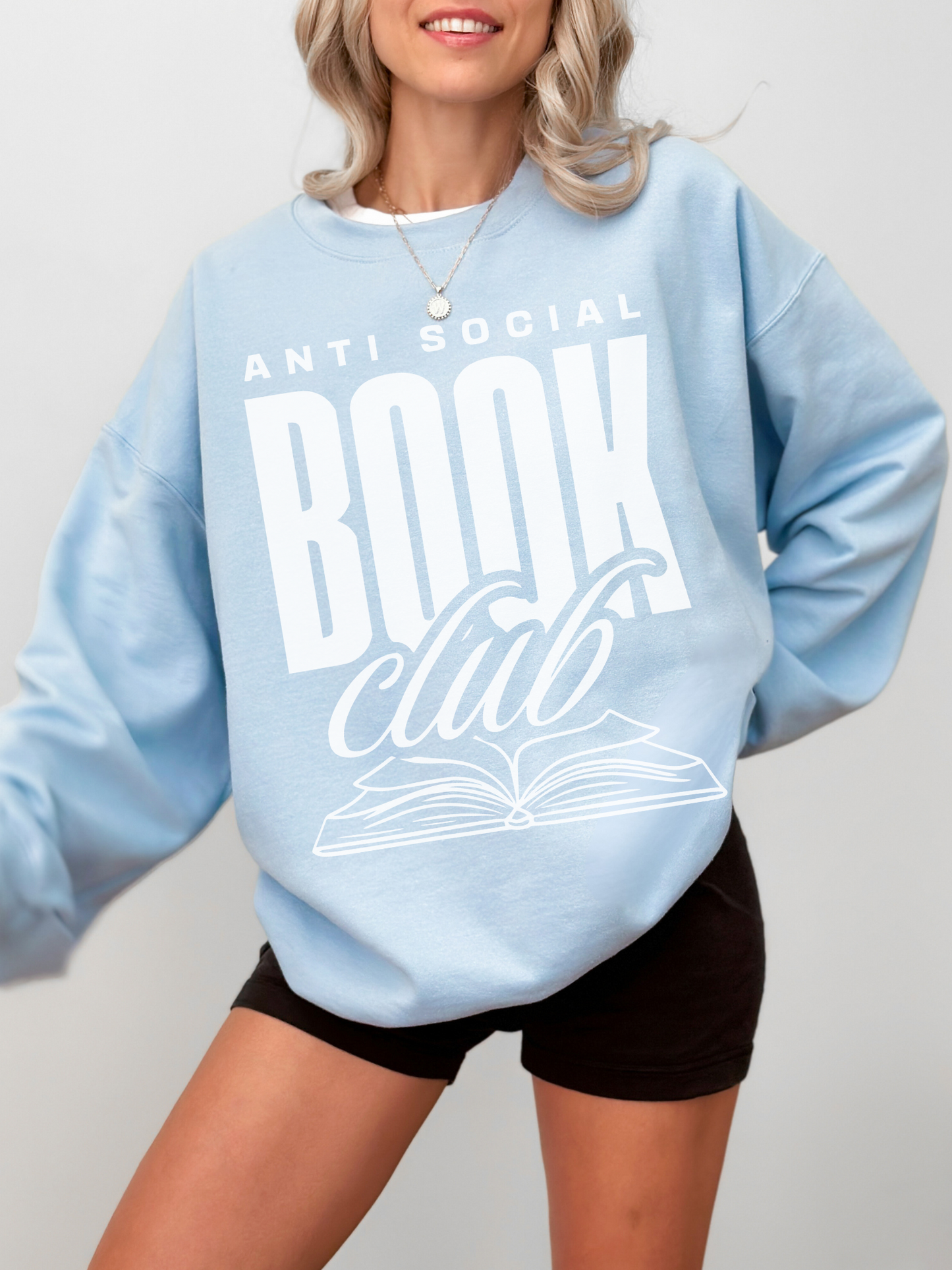 woman in pastel blue sweatshirt that says anti social book club in large white font with open book