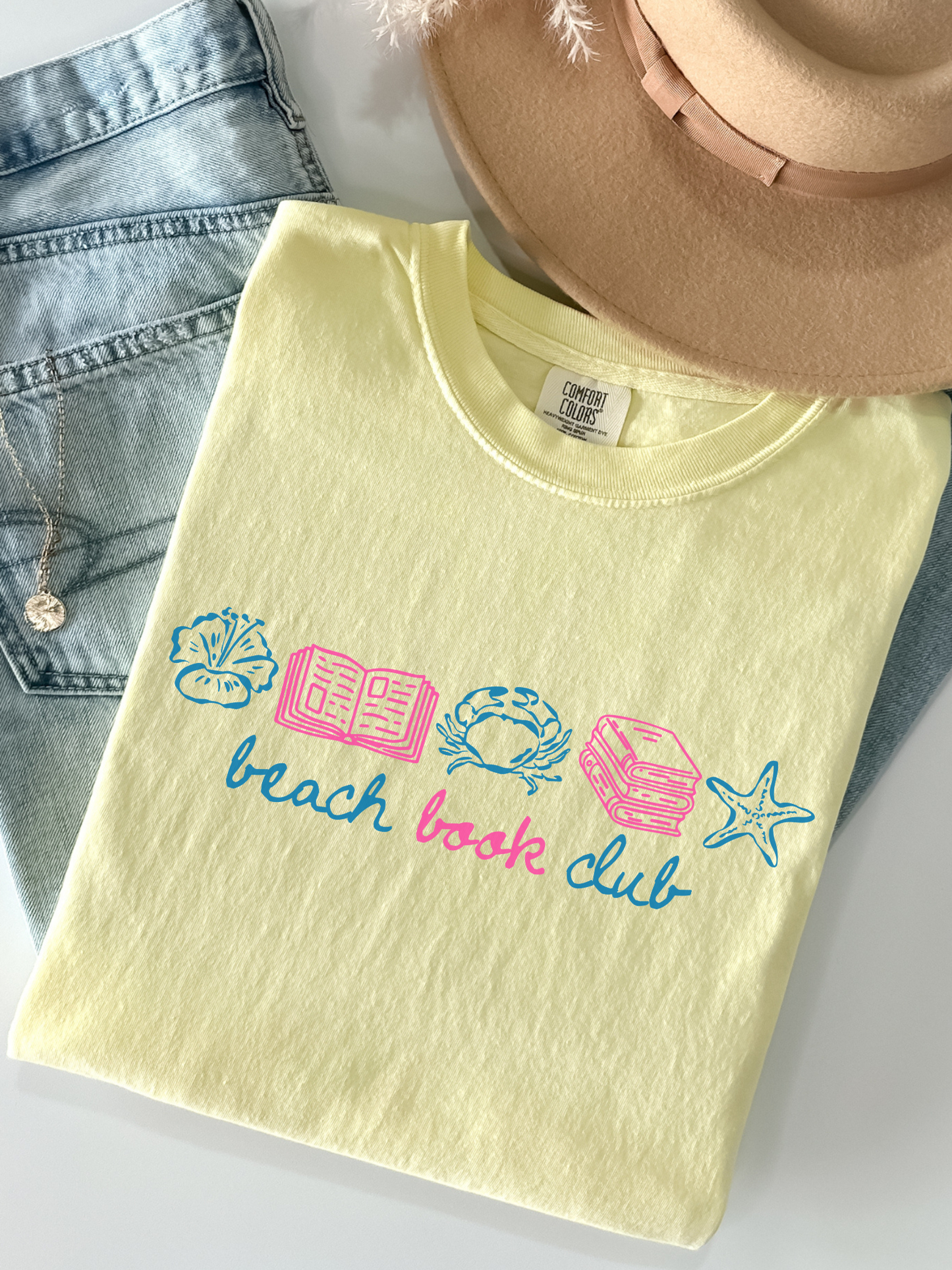 butter yellow comfort colors beach book club shirt flat lay with denim jeans hat necklace and colorful coastal reading graphic featuring seashell book crab books and starfish