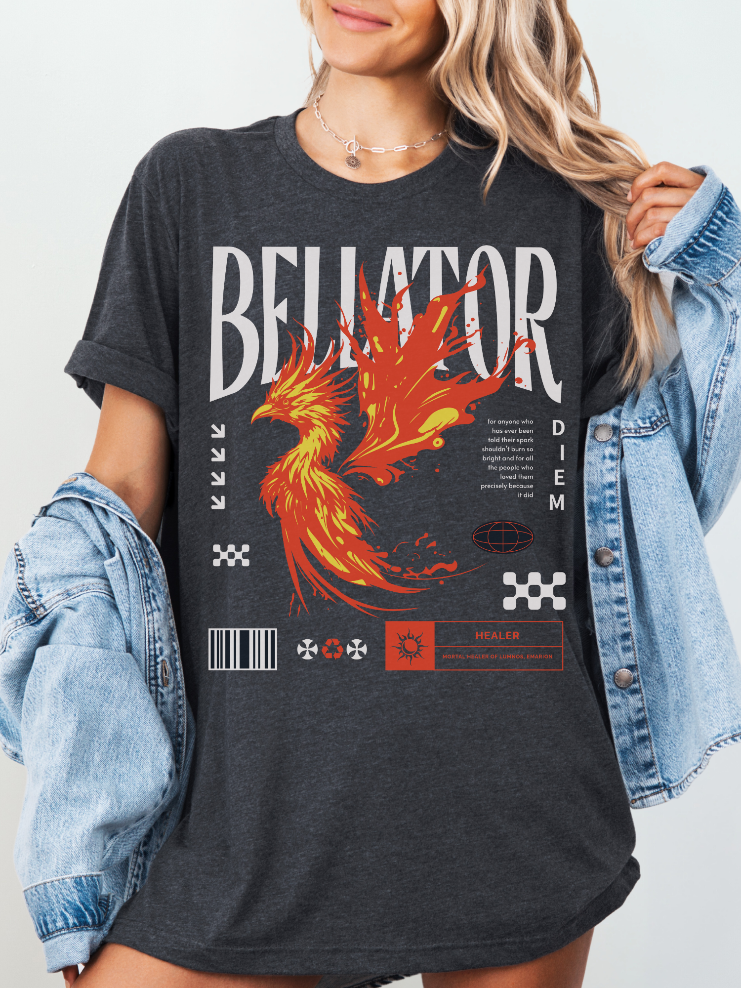 Woman in a dark grey heather Bellator phoenix shirt with rolled sleeves and denim jacket, showcasing a striking red and yellow flaming phoenix and fantasy text with barcode and emblem accents.