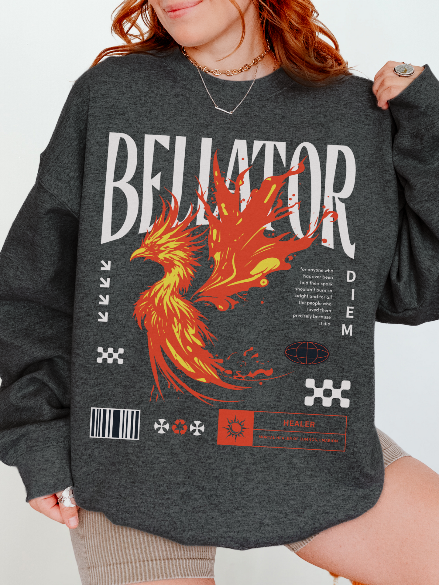 Dark heather grey sweatshirt styled with phoenix graphic and bold white Bellator lettering. Model with rolled sleeves and long blonde hair, layered with a denim jacket.