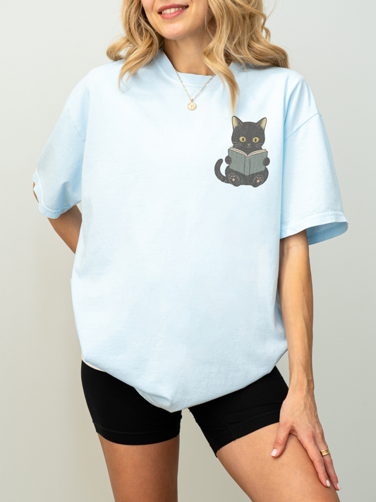 woman wearing a chambray Comfort Colors shirt featuring a black cat reading a book chest graphic, cozy reader shirt for book lovers and cat moms