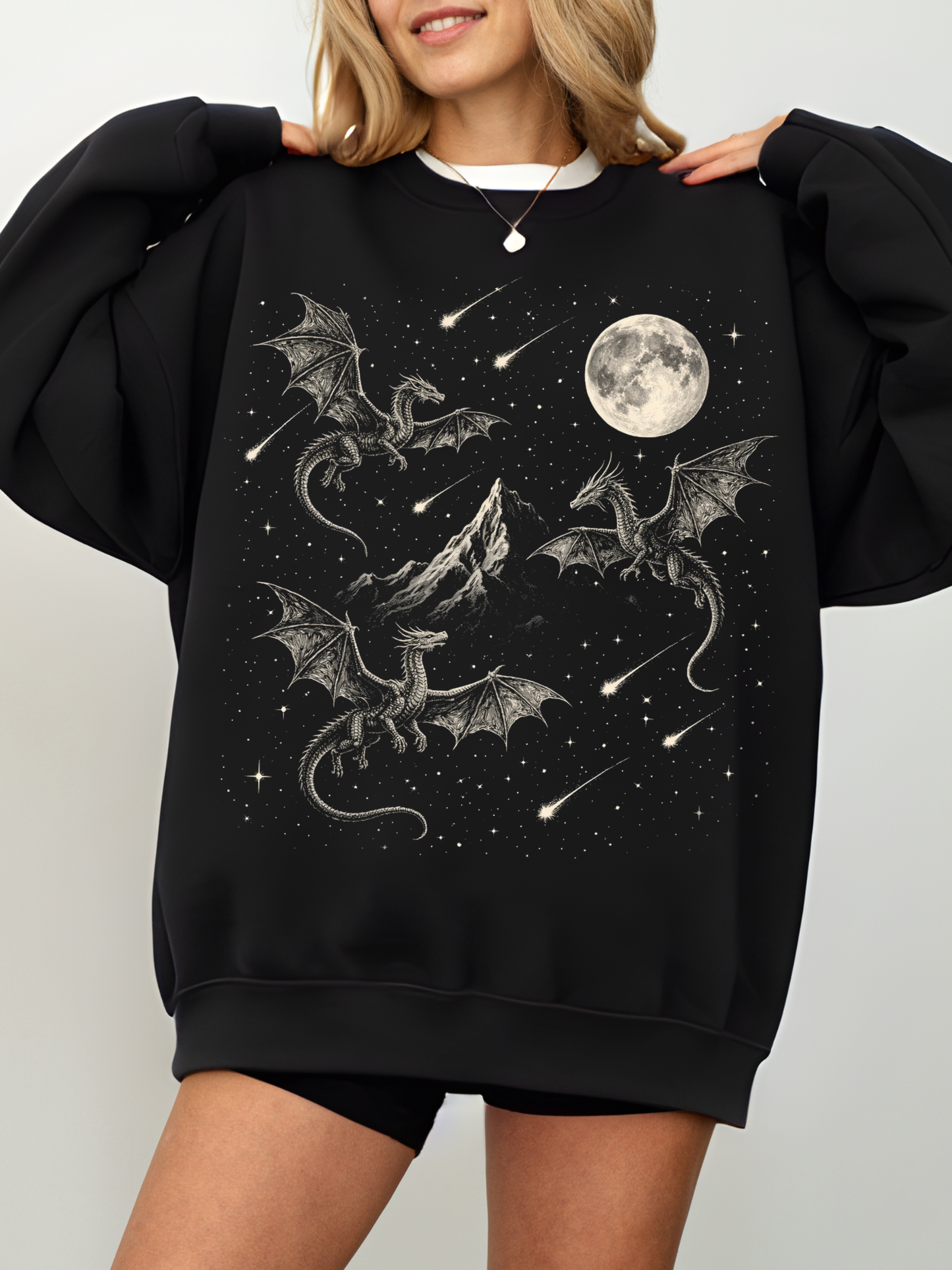 Black oversized sweatshirt featuring a detailed fantasy dragon scene with multiple dragons flying around a mountain under a full moon and starry night sky with shooting stars. Cozy fantasy sweatshirt for dragon lovers, readers, and dark aesthetic outfits.