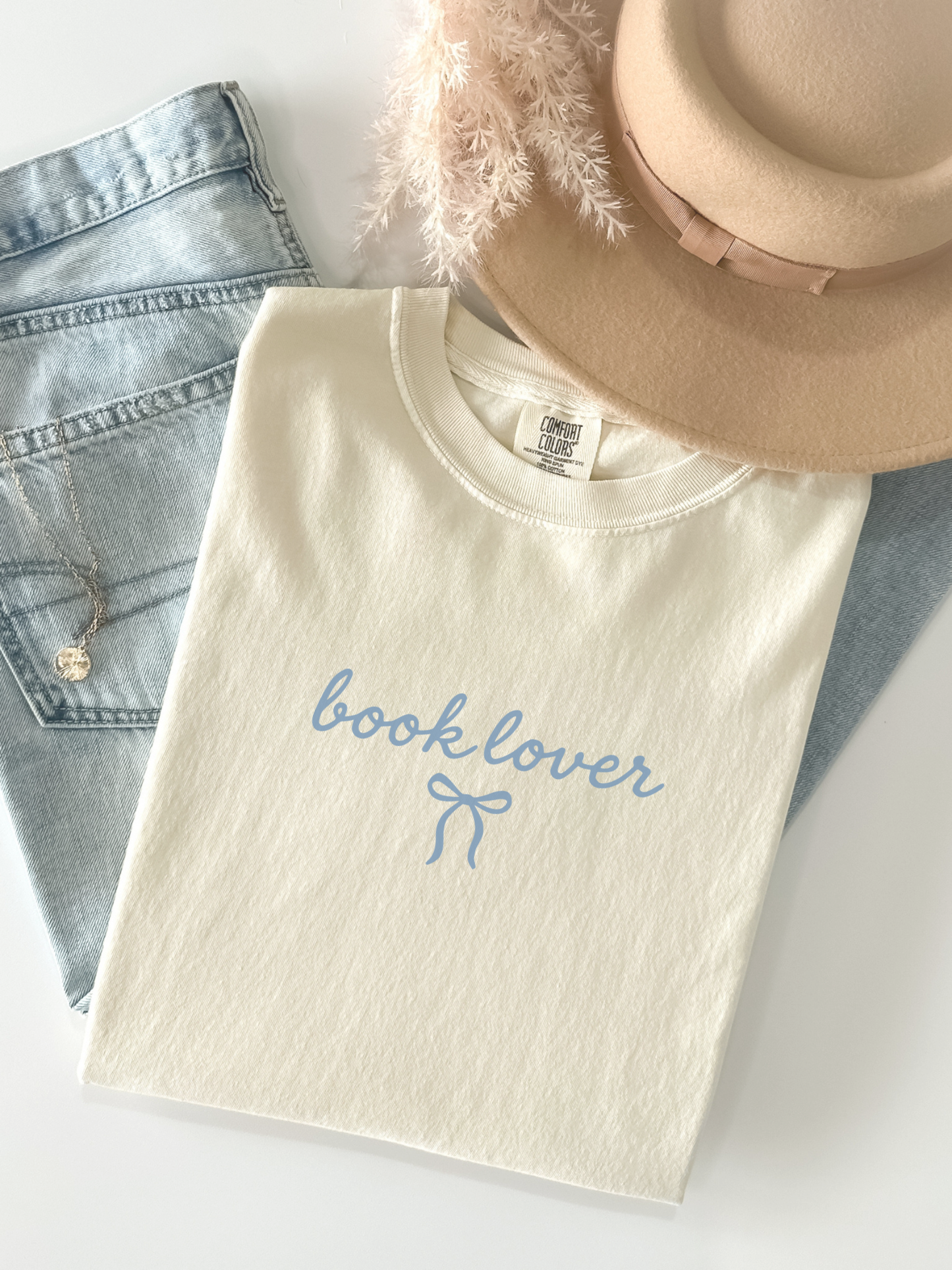 flat lay of cream book lover shirt with bow next to hat and jeans