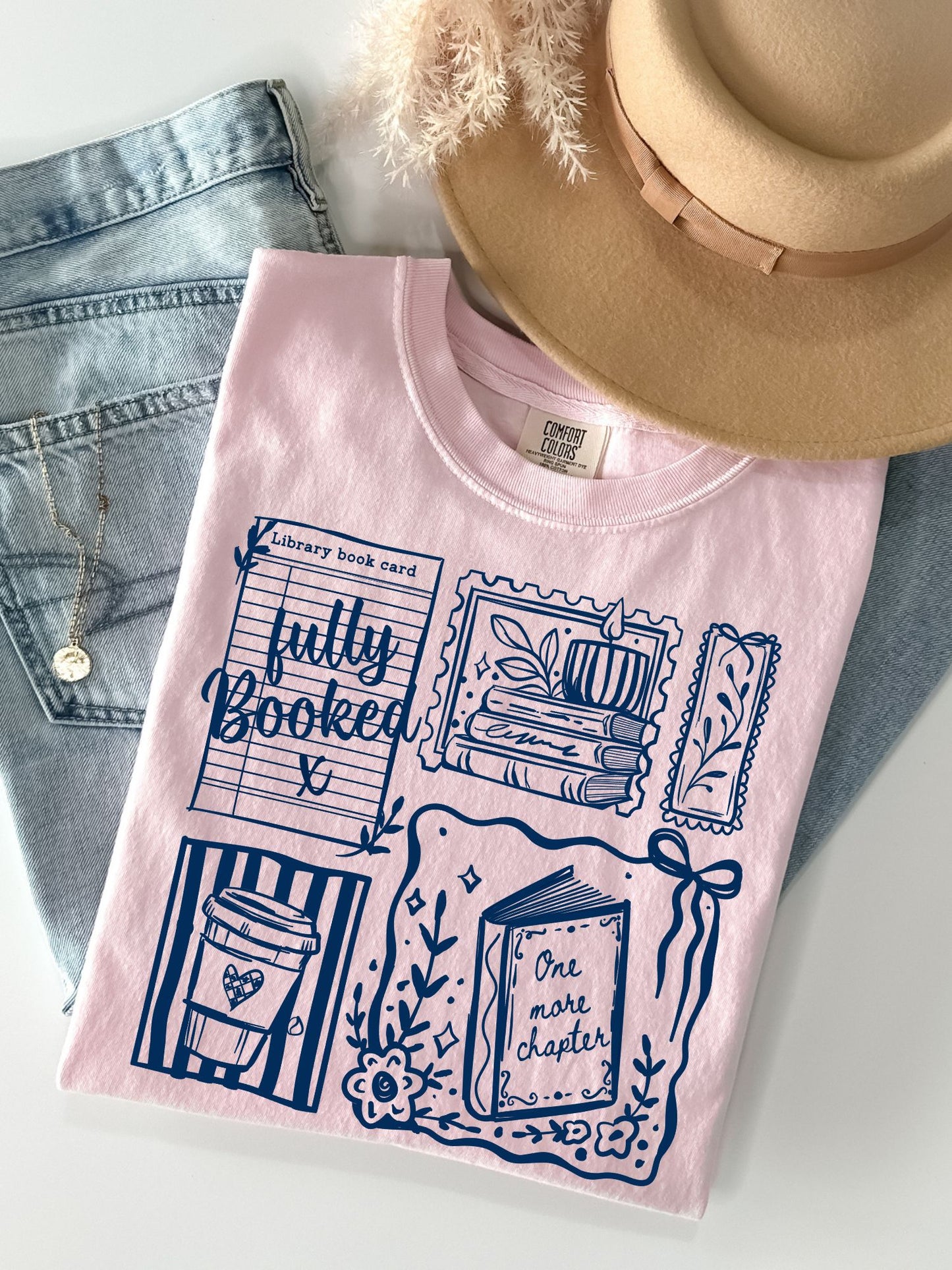 A light pink graphic T-shirt laid flat on a white surface, styled with blue jeans, a tan hat, and a necklace. The shirt has a navy blue book-themed print with phrases like 'fully Booked' and 'One more chapter.