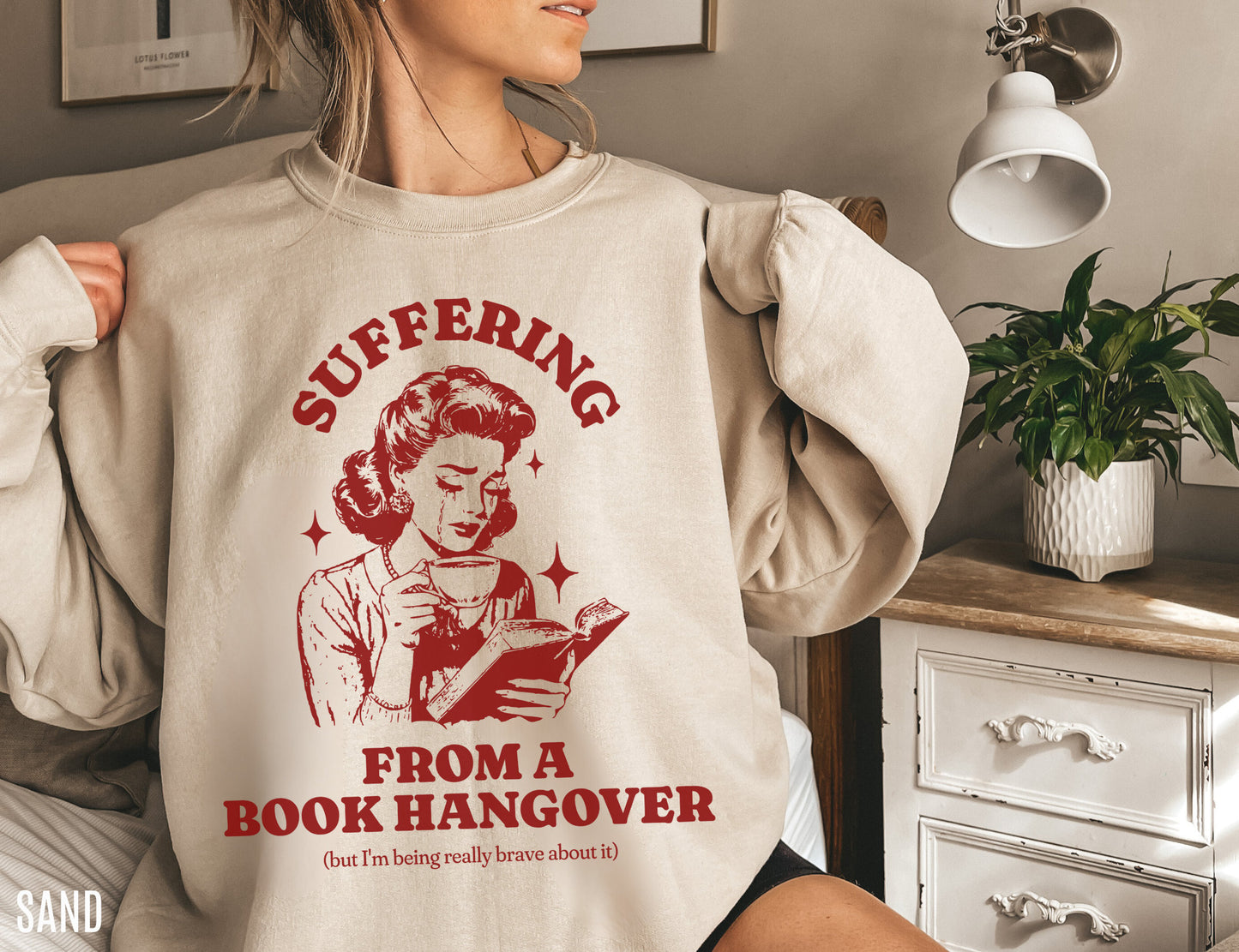 woman lounging in sand-colored sweatshirt that says suffering from a book hangover in red retro font