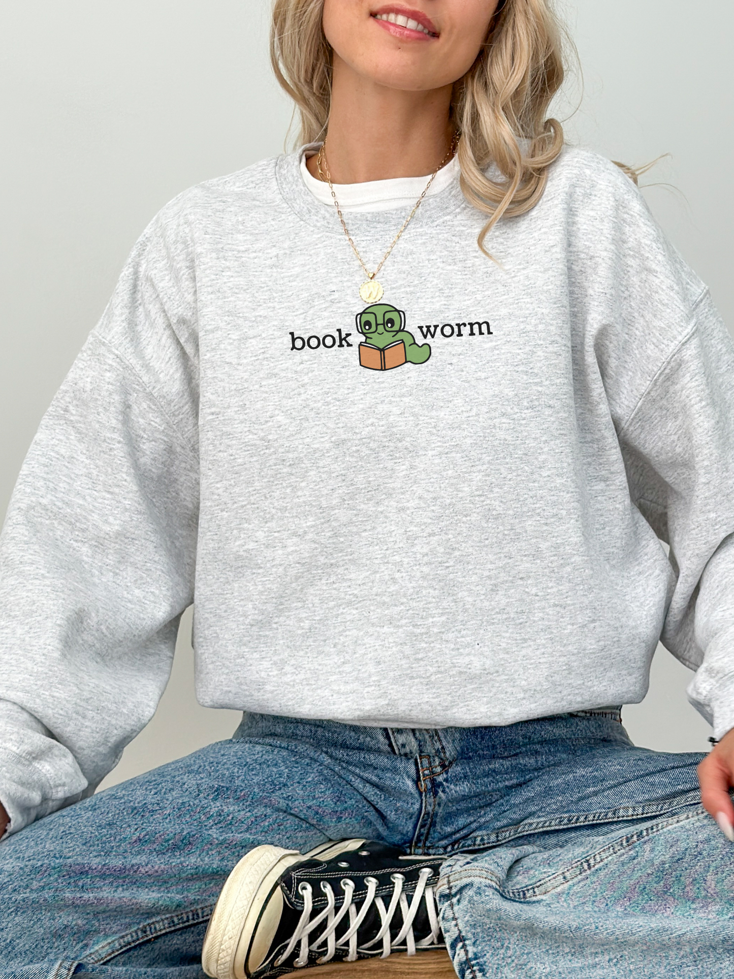 woman sitting cross-legged in an ash grey sweatshirt with a book worm design featuring a cute green worm and book