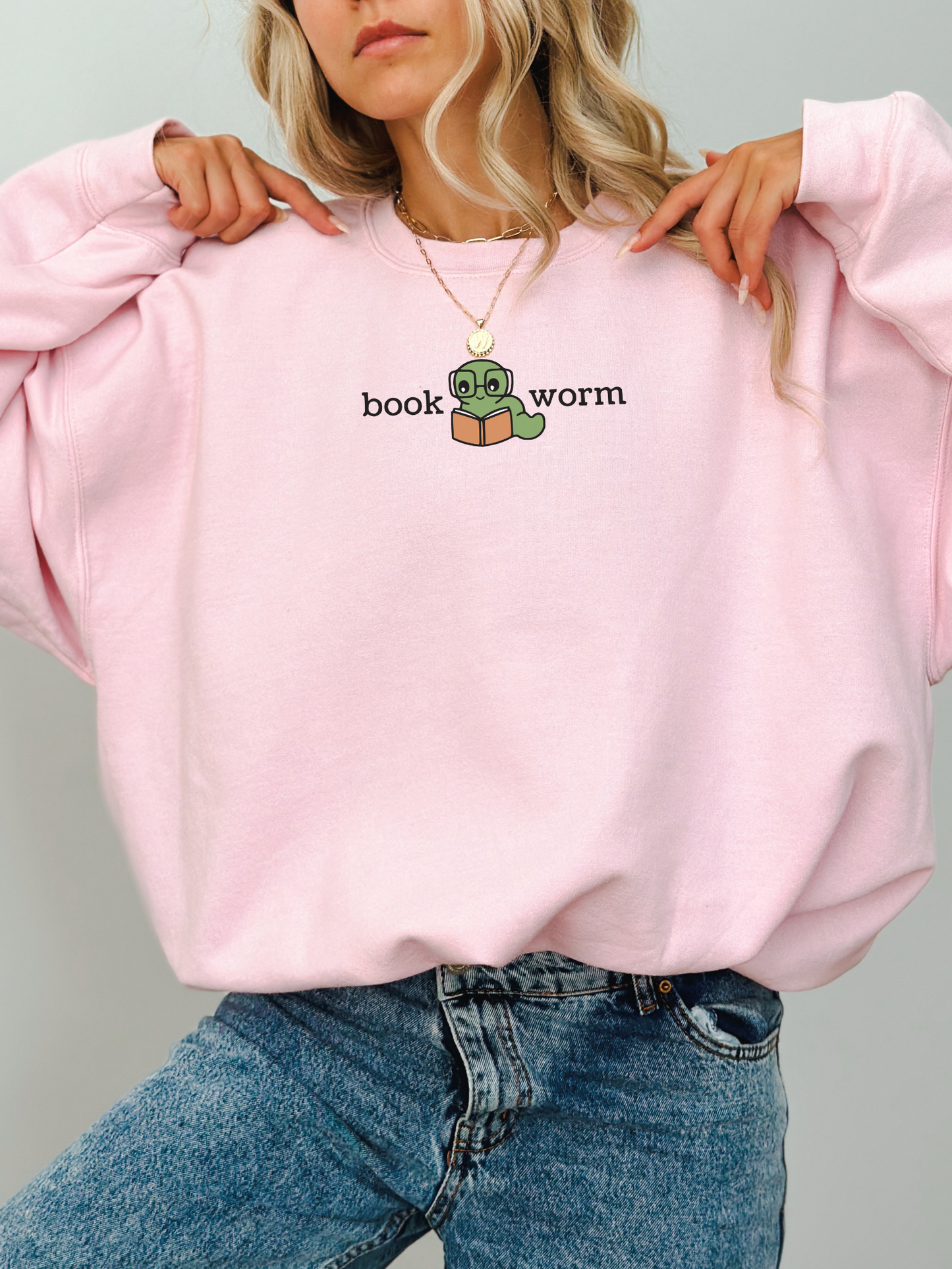 woman wearing a pink sweatshirt that says book worm with a cute green worm in glasses reading a book