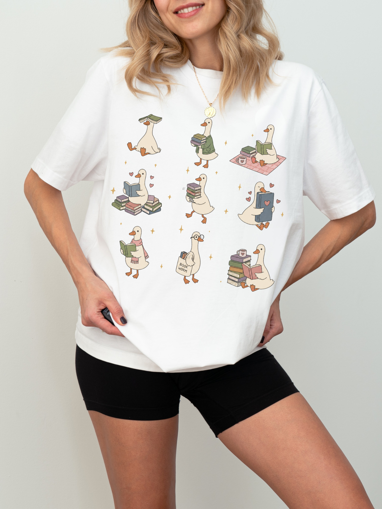 woman wearing white comfort colors book lover goose shirt featuring cute reading ducks stacks of books coffee mugs and cozy reader design
