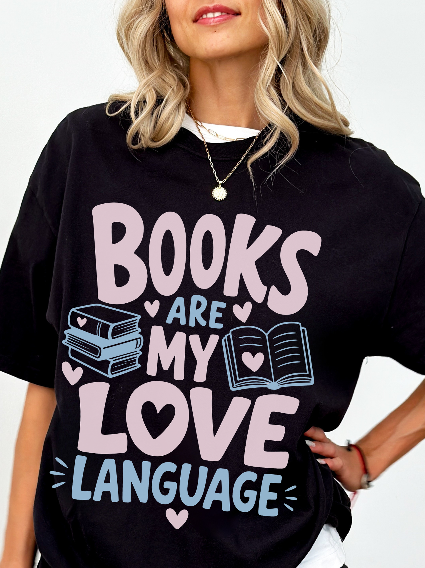 woman wearing a black comfort colors t shirt featuring books are my love language graphic with pastel pink and blue text stacked books open book illustration and heart details for book lovers