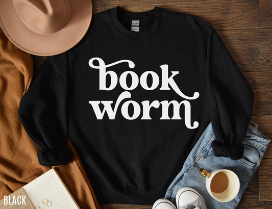 Black bookworm sweatshirt with large white text laid flat with fall accessories including a hat, cardigan, book, and coffee