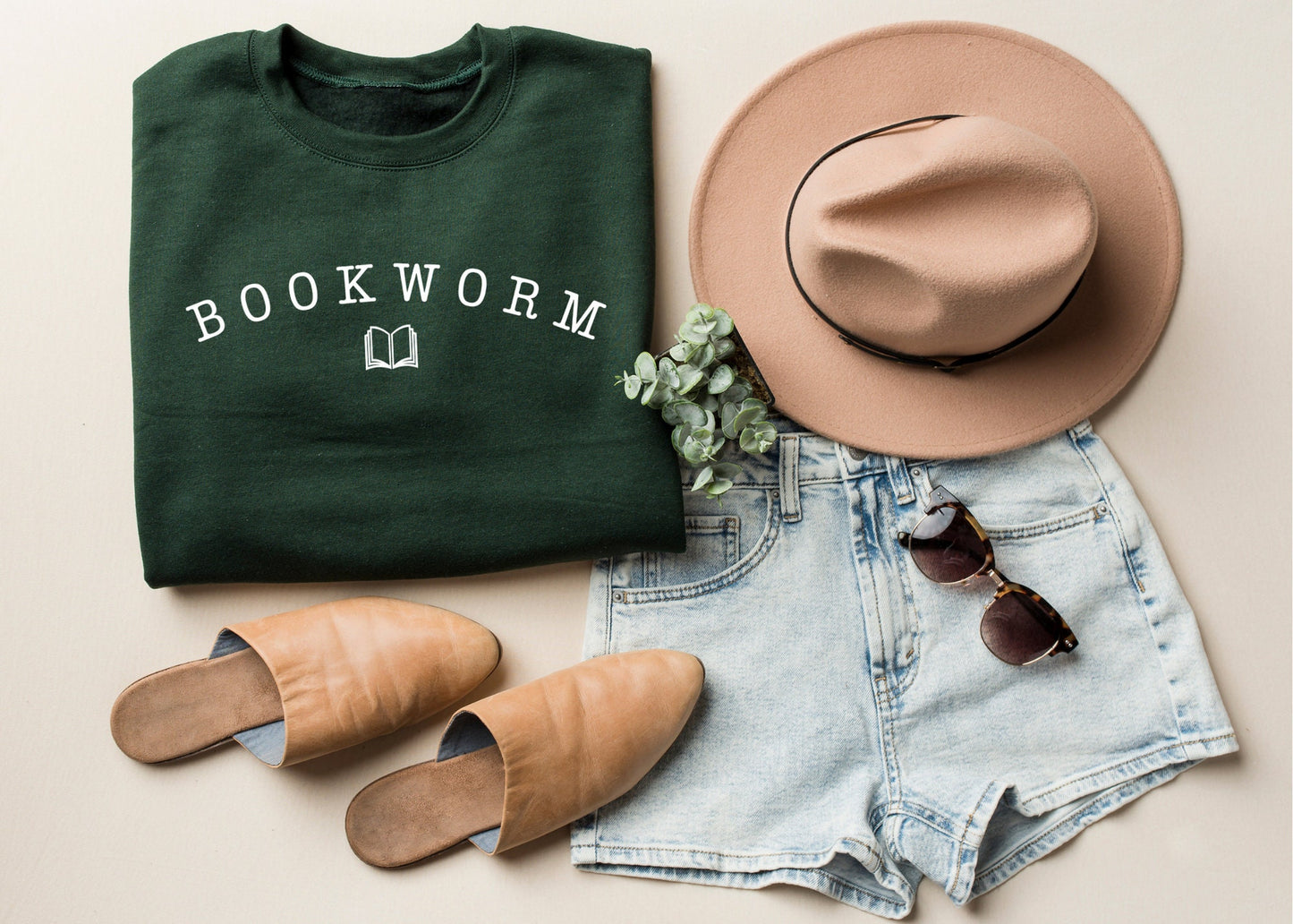 forest green bookworm sweatshirt folded with white text and book icon, styled with tan hat, sandals, and denim shorts