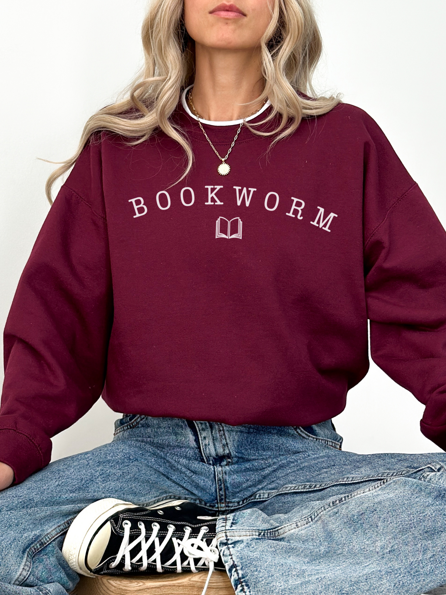 woman wearing a maroon bookworm sweatshirt with white collegiate lettering and book icon, styled with layered gold necklaces and jeans