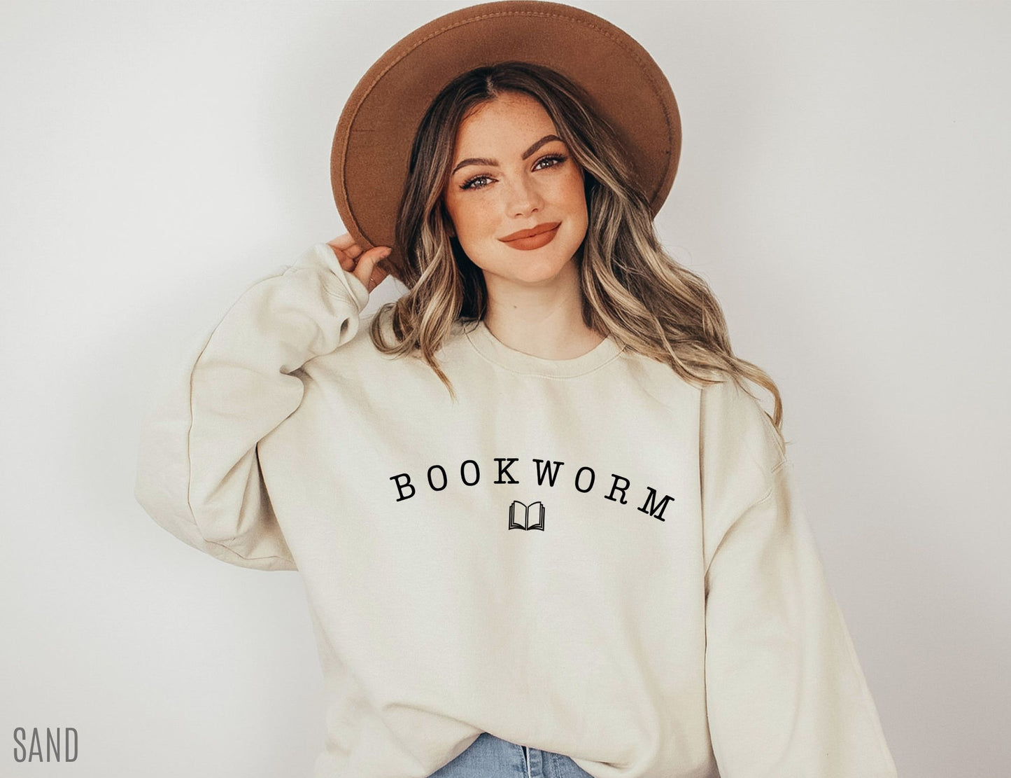 woman wearing a sand-colored bookworm sweatshirt with curved black text and open book icon, styled with gold chain necklace and denim jeans
