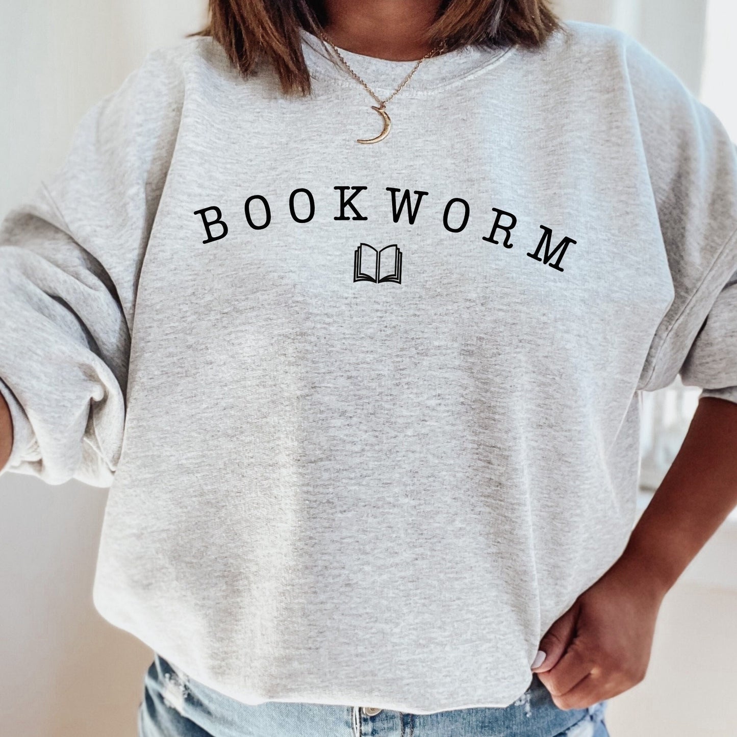 woman wearing a sport grey bookworm sweatshirt with black arched lettering and open book symbol, seated with plant decor in the background