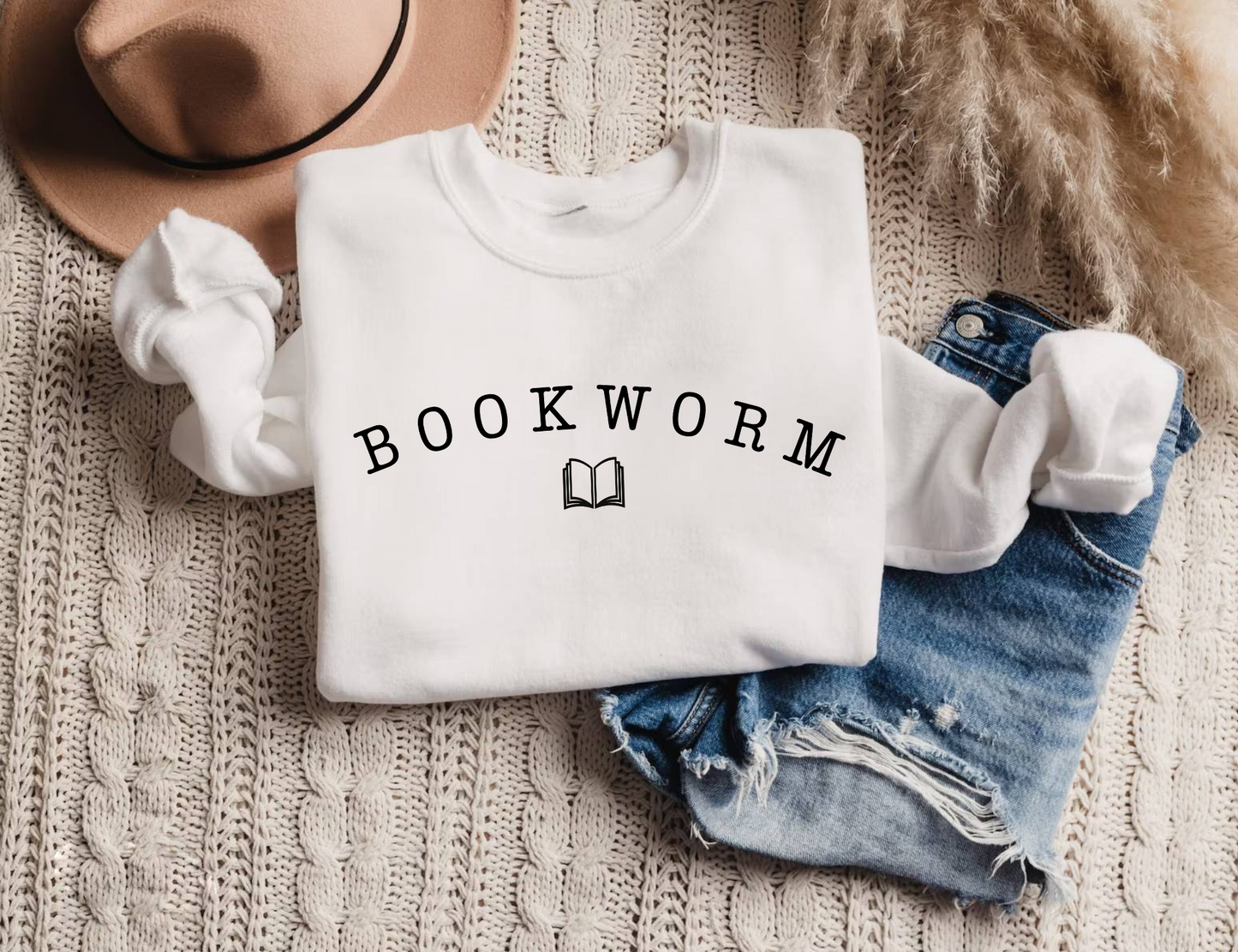 white bookworm sweatshirt folded with black varsity-style text and book graphic, styled with jeans, coffee, and a tan hat
