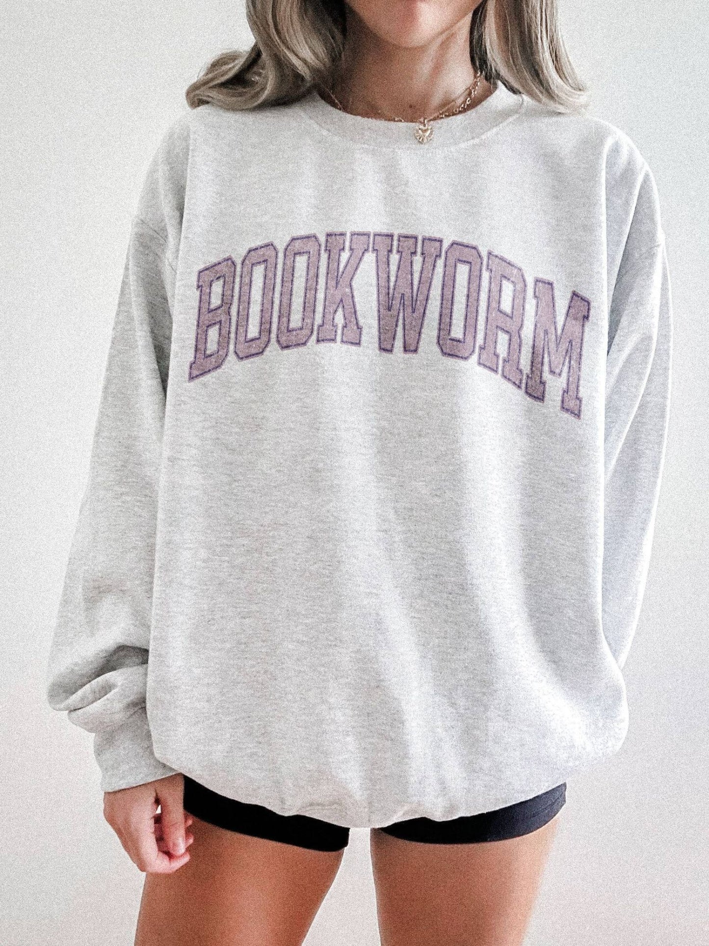 girl standing wearing an ash grey sweatshirt that says "bookworm" across the top in a jersey sport font