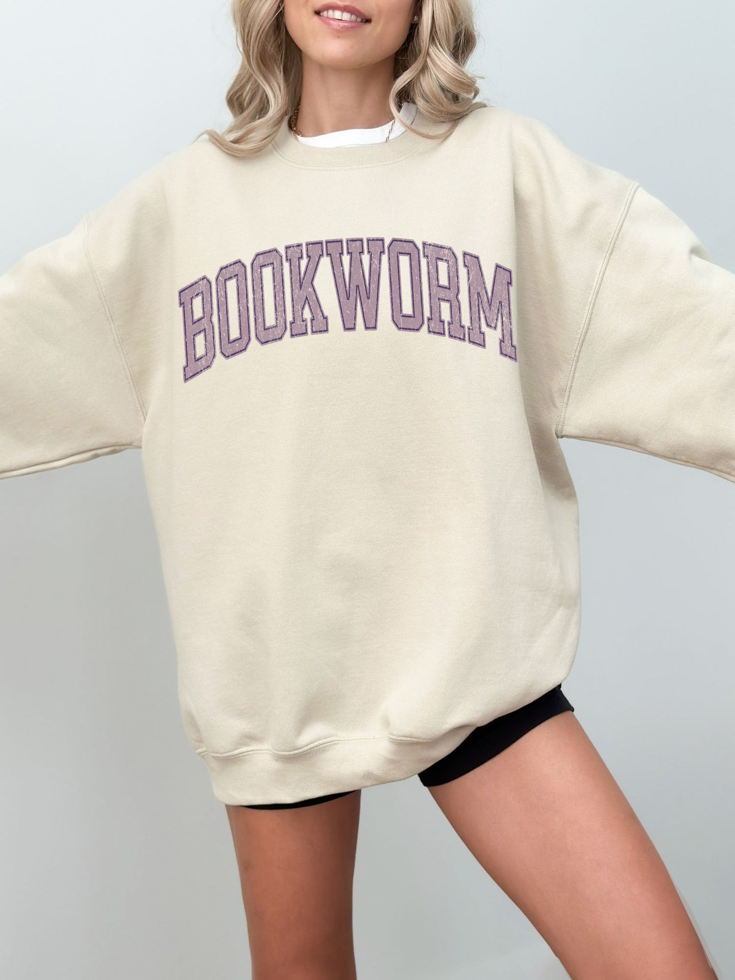 girl standing wearing a sand sweatshirt that says "bookworm" across the top in a purple jersey sport font