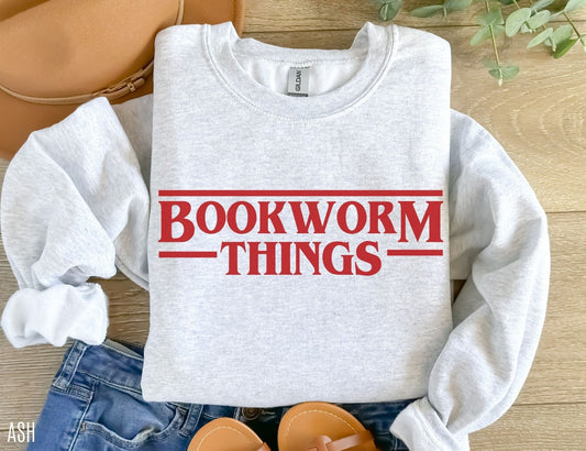 folded ash grey sweatshirt with red bookworm things text, styled with jeans, sandals, and greenery