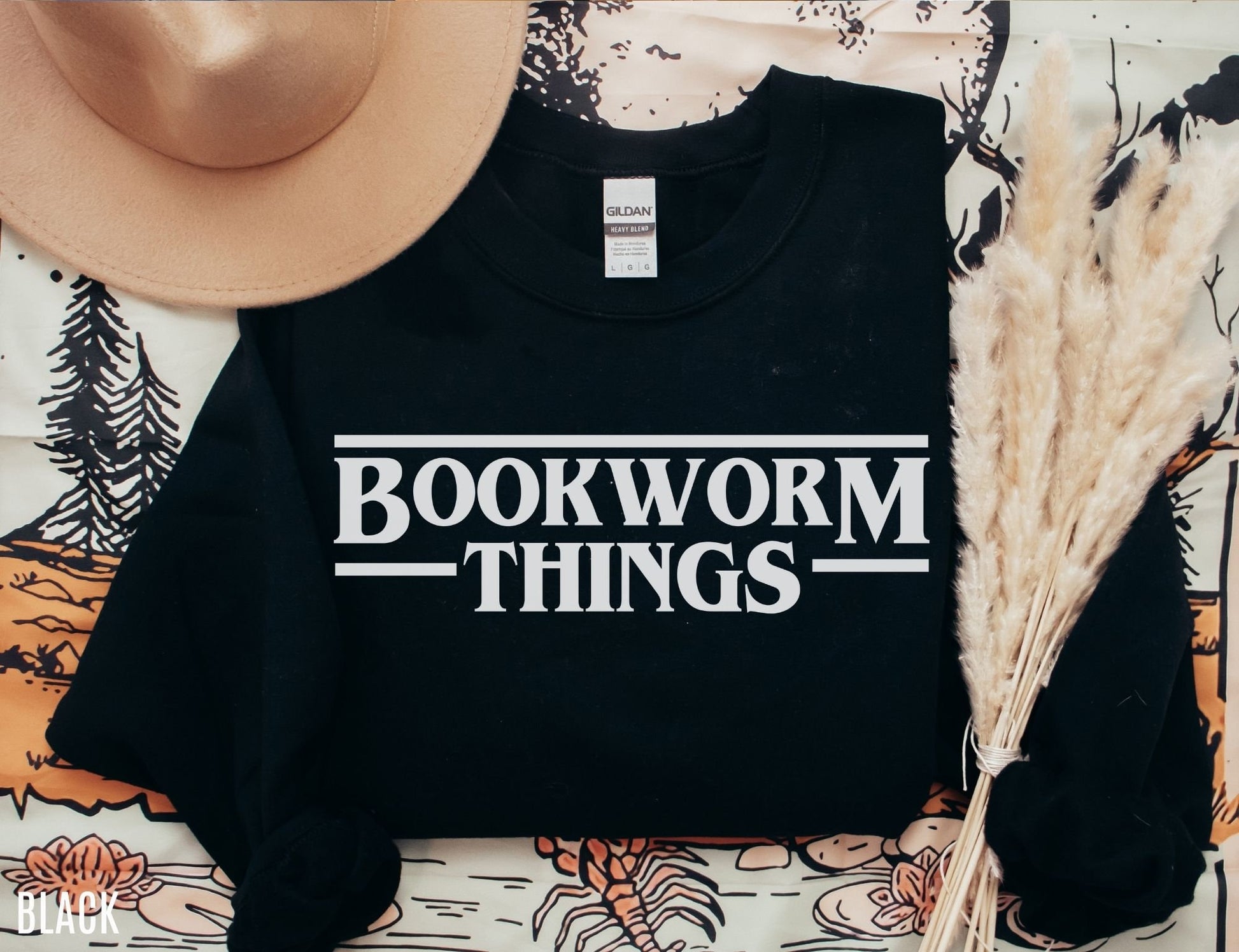 folded black sweatshirt with white bookworm things design laid out with pampas grass and hat