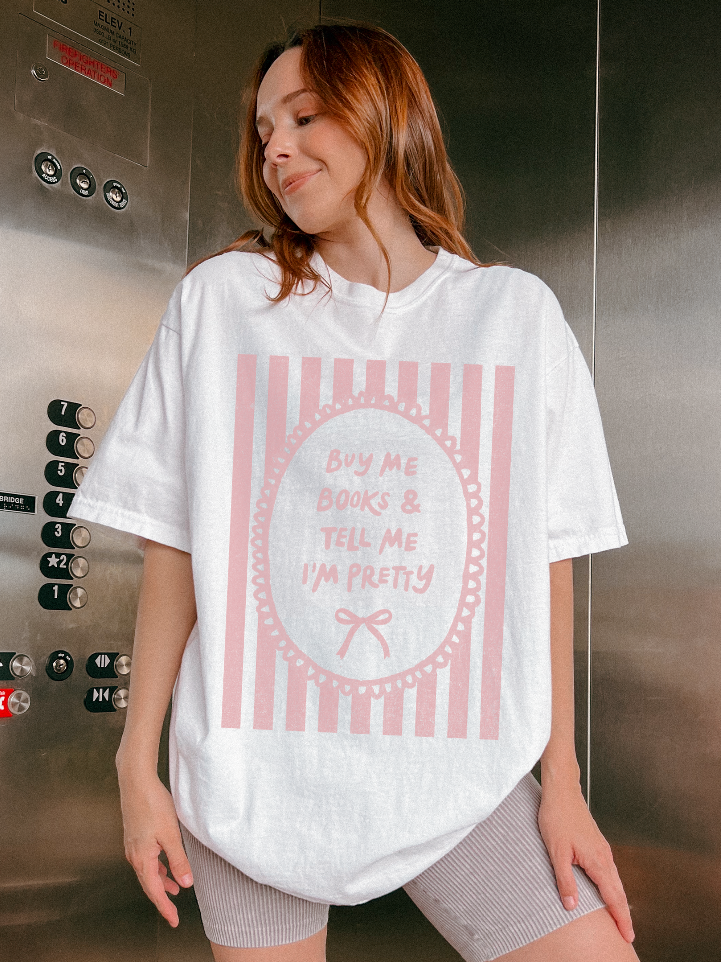 Woman wearing a cream oversized t shirt with pink striped vintage frame graphic and text buy me books and tell me I’m pretty styled casually indoors