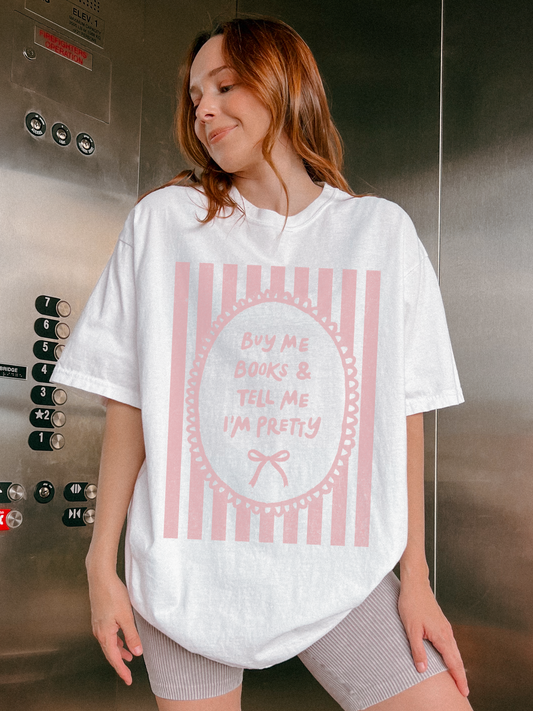 Woman wearing a cream oversized t shirt with pink striped vintage frame graphic and text buy me books and tell me I’m pretty styled casually indoors
