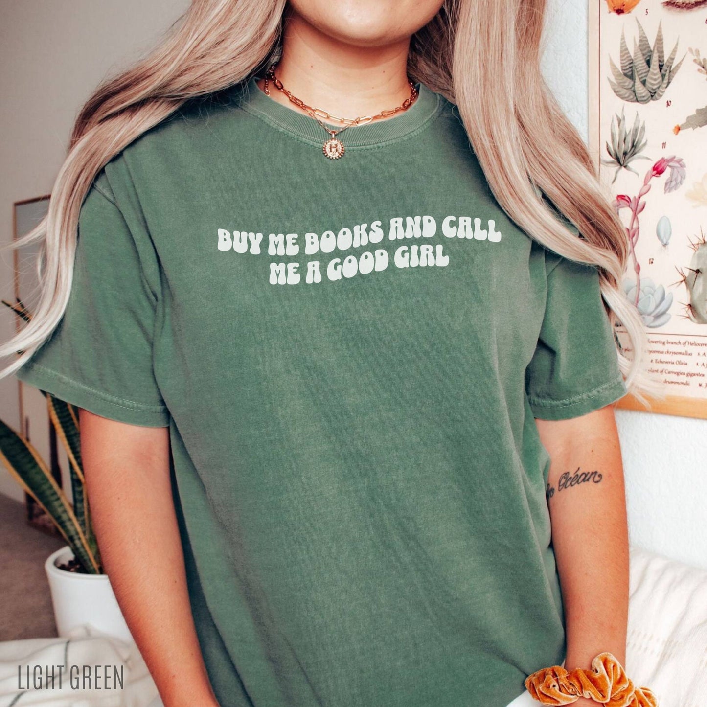 woman wearing light green shirt that says buy me books and call me a good girl standing in front of plant print wall art
