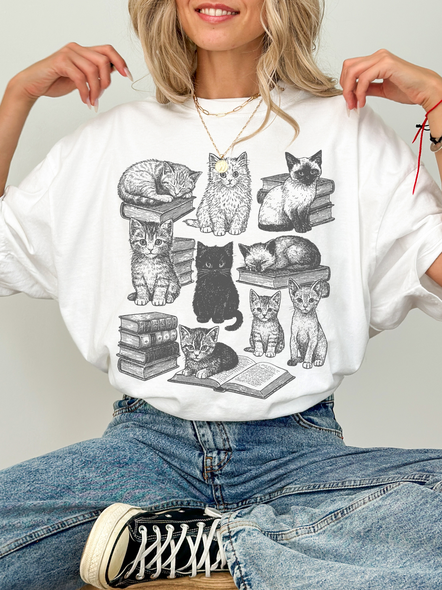 woman sitting cross-legged wearing a white t-shirt with vintage sketch-style kittens and cats sitting on books, cozy aesthetic for book lovers and cat fans