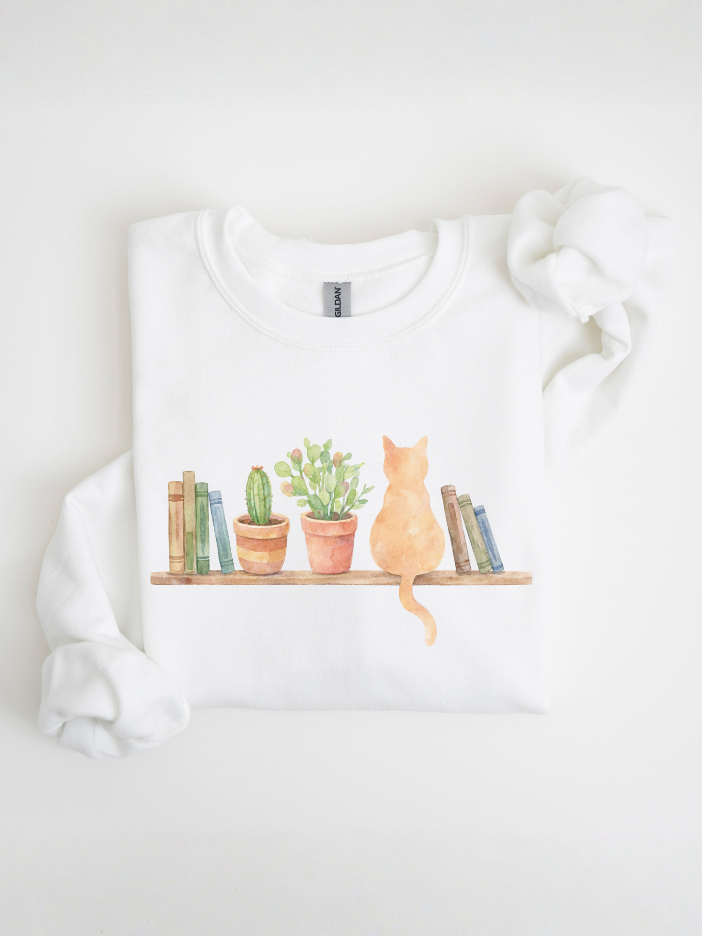 folded white sweatshirt with watercolor cat sitting among books and plants displayed on a clean background