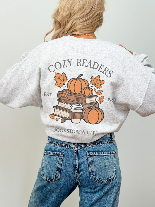 Back view of light grey Cozy Readers sweatshirt showing large graphic of stacked books, pumpkins, fall leaves, and a takeaway coffee cup with the text Cozy Readers Bookstore and Cafe EST 1983.