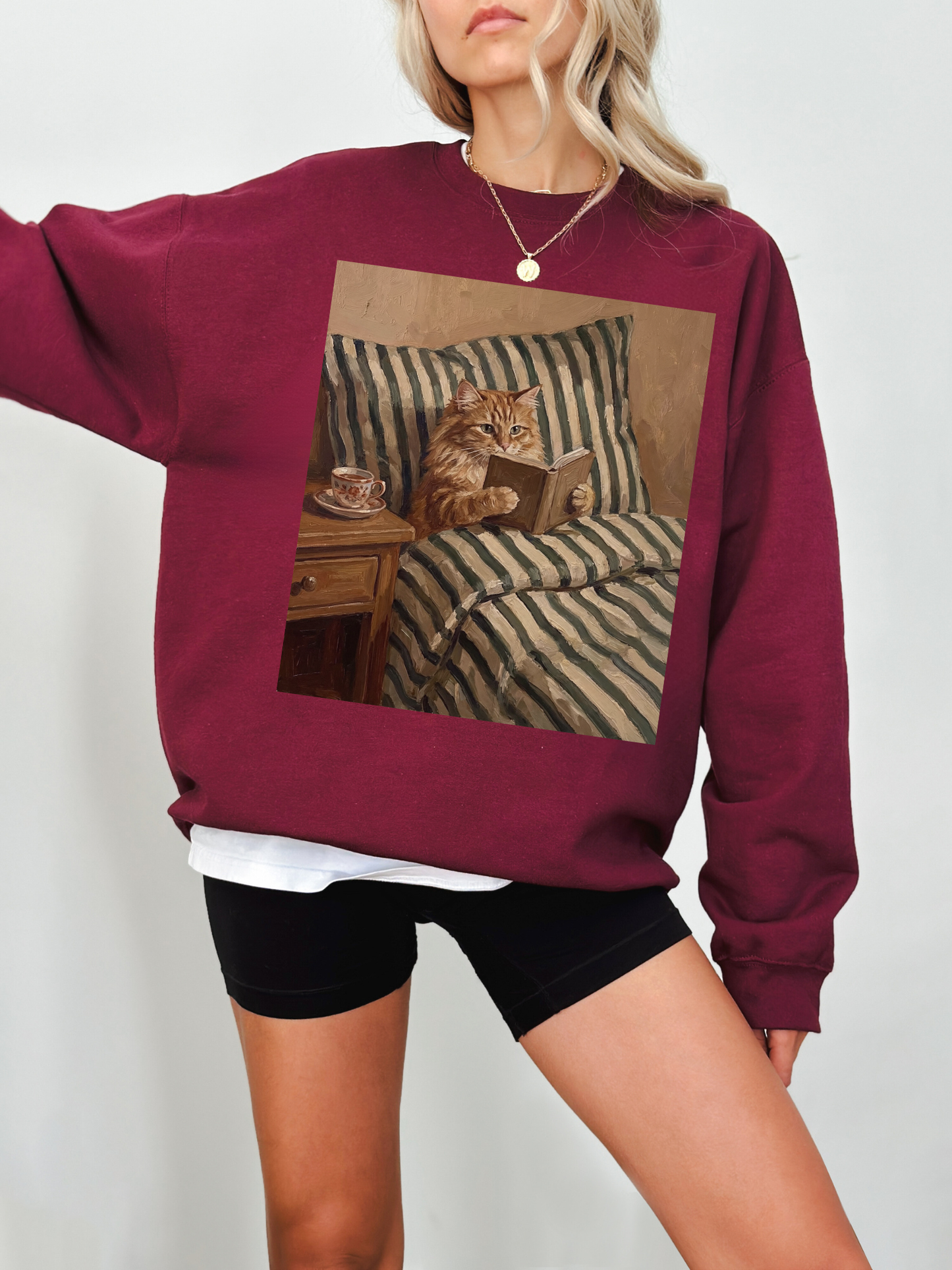 Maroon sweatshirt featuring a cozy vintage style cat reading a book in bed with a bedside table and tea cup, worn by a model indoors. The rich color and relaxed fit create a warm and inviting aesthetic, perfect for book lovers, readers, and anyone who loves cozy cat themed clothing.