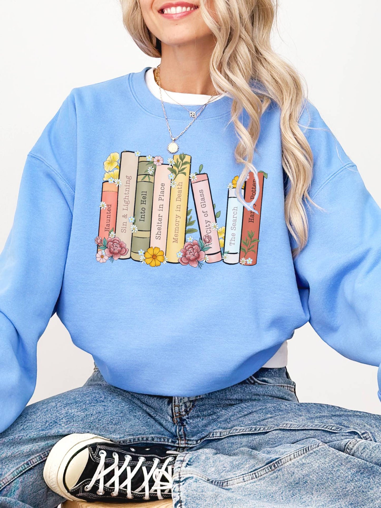 woman wearing light blue sweatshirt with colorful book stack including Haunting Adeline, Sky Without Stars, and Divergent