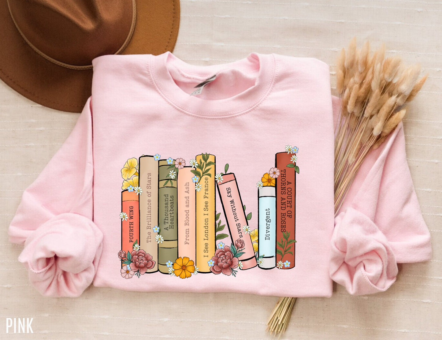 folded pink sweatshirt with pastel book spine design featuring floral fantasy titles like ACOTAR and Fourth Wing