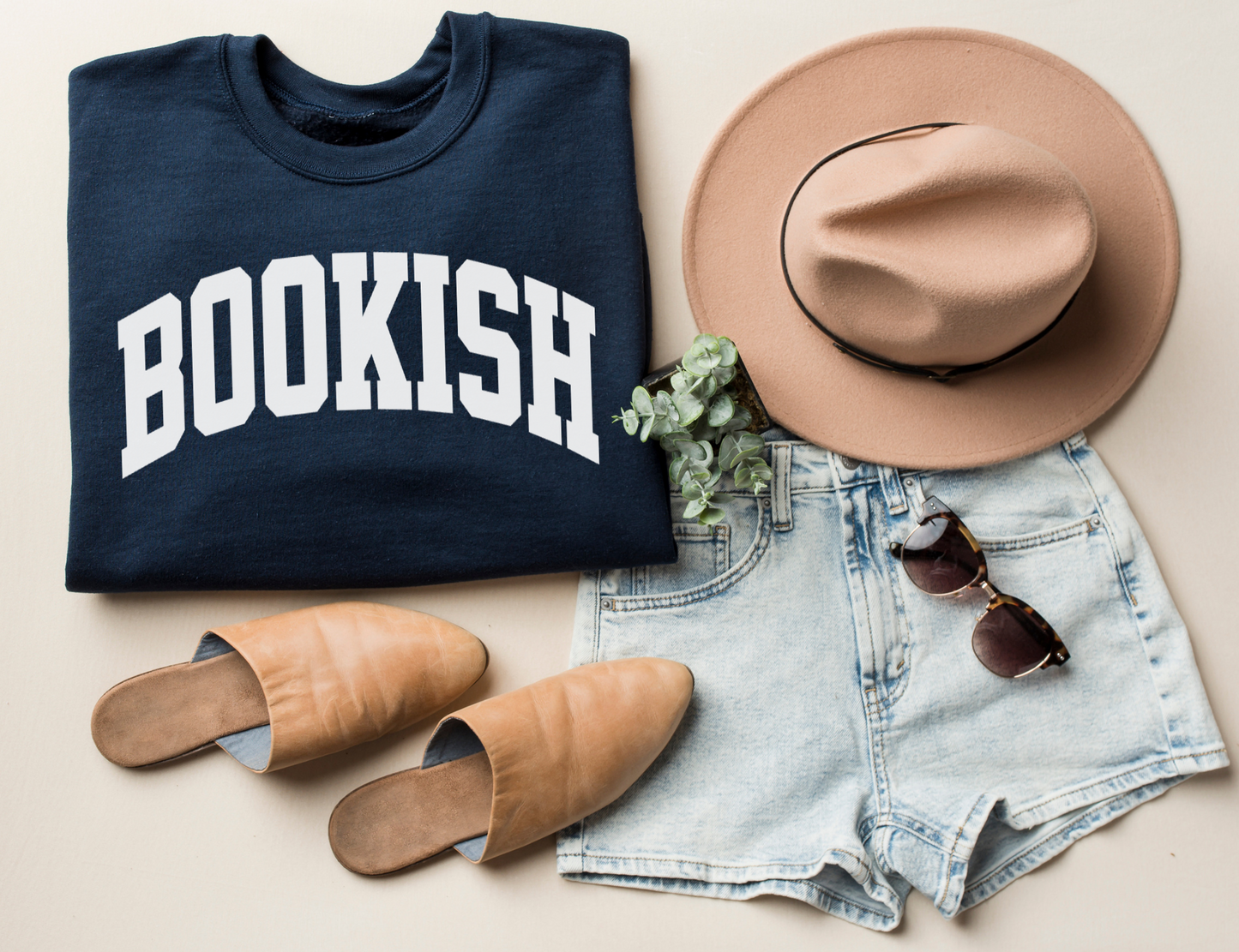 folded navy sweatshirt with bold white varsity-style BOOKISH text, styled with tan hat, sunglasses, and denim shorts
