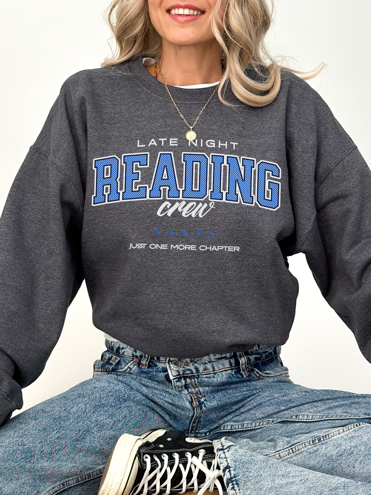 Woman wearing dark heather grey bookish sweatshirt that says “Late Night Reading Crew” in blue varsity font with denim jeans.