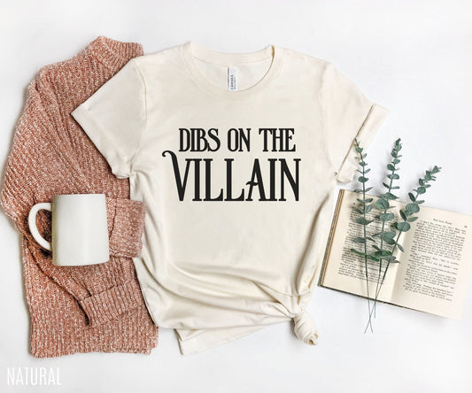 flat lay of natural color Dibs on the Villain t-shirt with book, sweater, and greenery