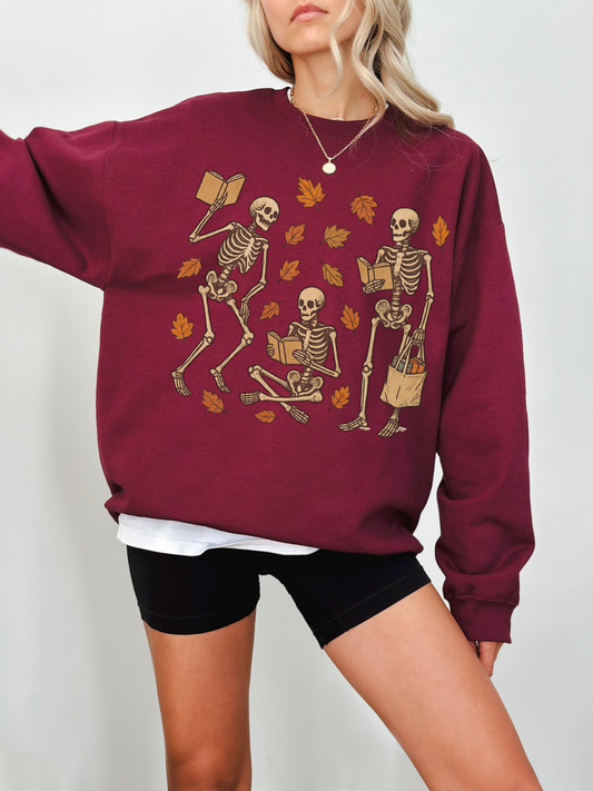 Woman wearing maroon Gildan sweatshirt with three reading skeletons and autumn leaves printed on the front, paired with black biker shorts and layered white tee