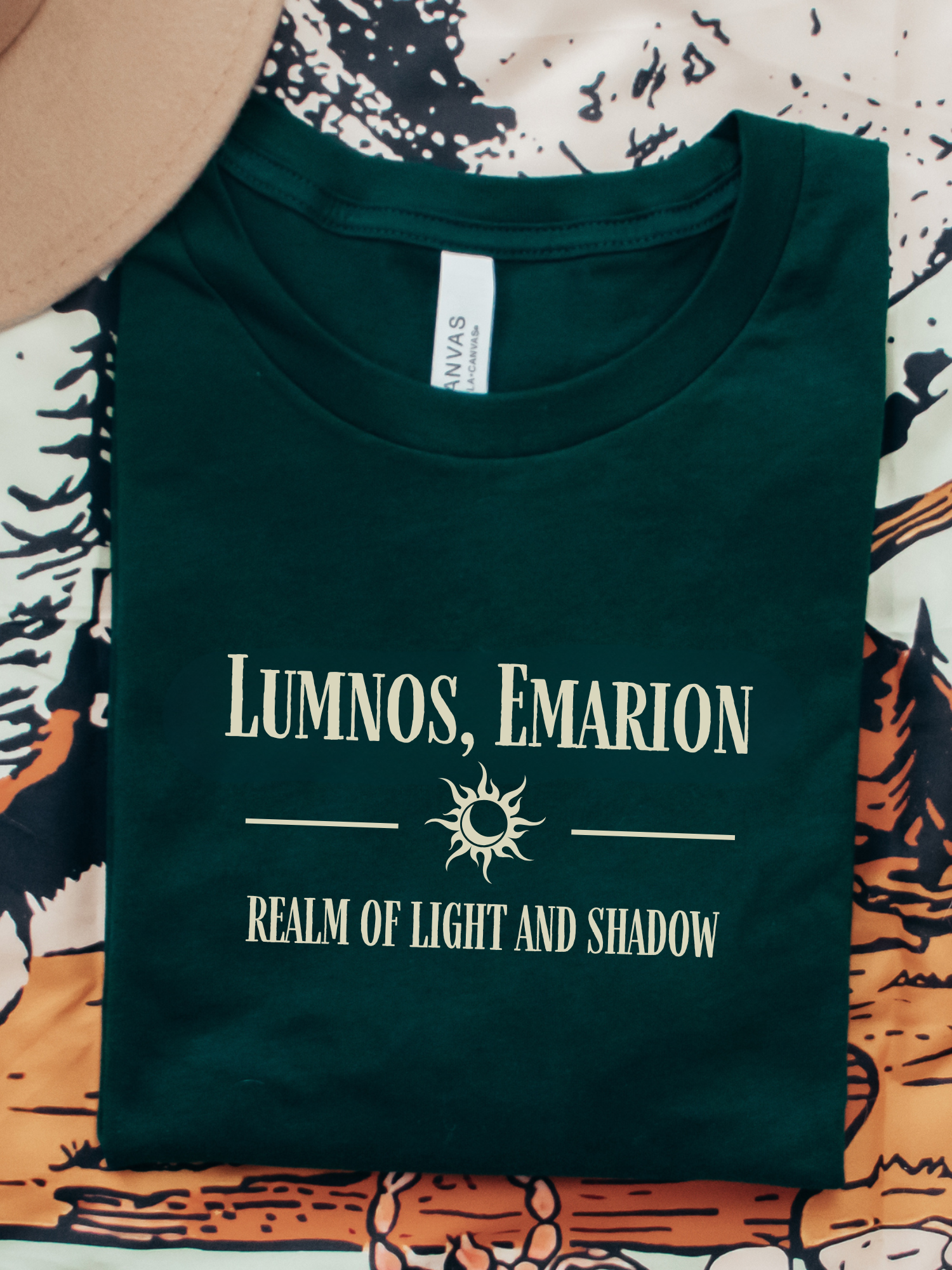 Flatlay of a forest green Bella Canvas shirt with cream fantasy quote design "Lumnos, Emarion - Realm of Light and Shadow", styled with a tan hat and fantasy-themed bookish background