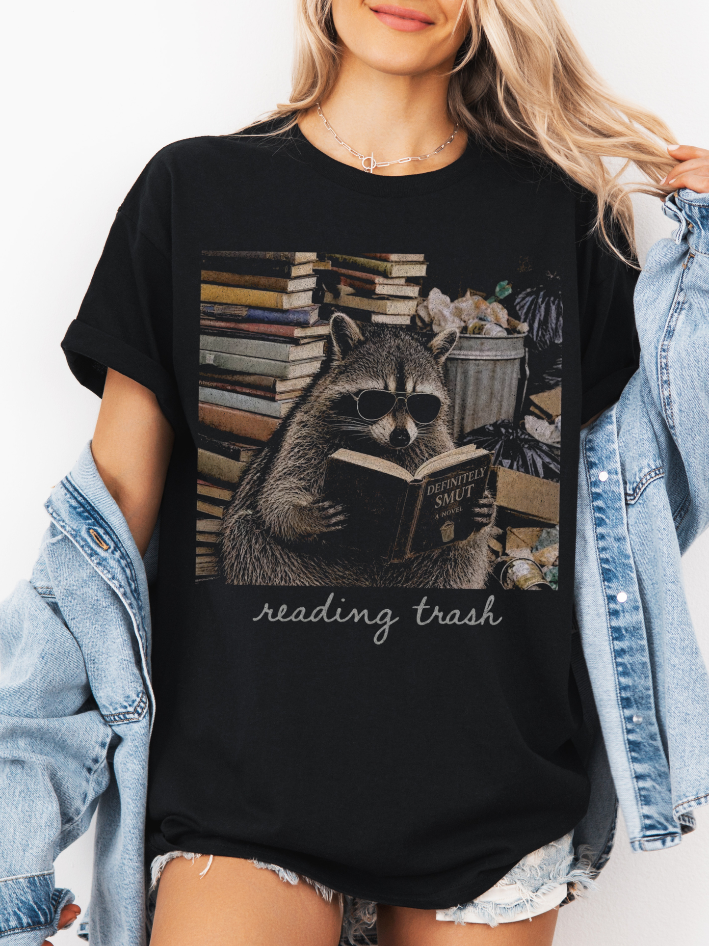 woman wearing black bookish raccoon shirt featuring reading trash graphic with sunglasses raccoon reading smut novel and denim jacket