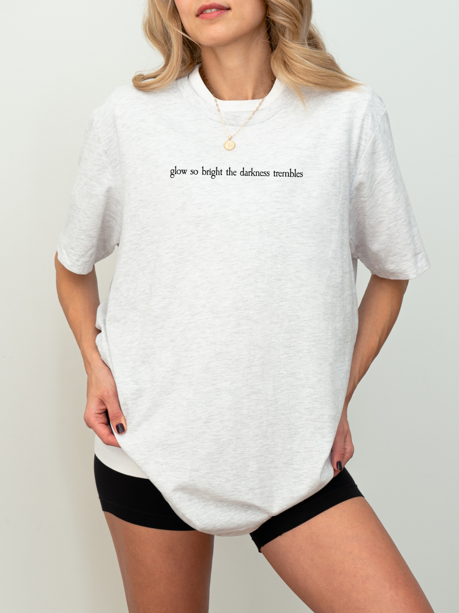 Woman modeling an ash gray Bella Canvas t-shirt with black serif text reading “glow so bright the darkness trembles,” paired with black shorts and a gold pendant necklace