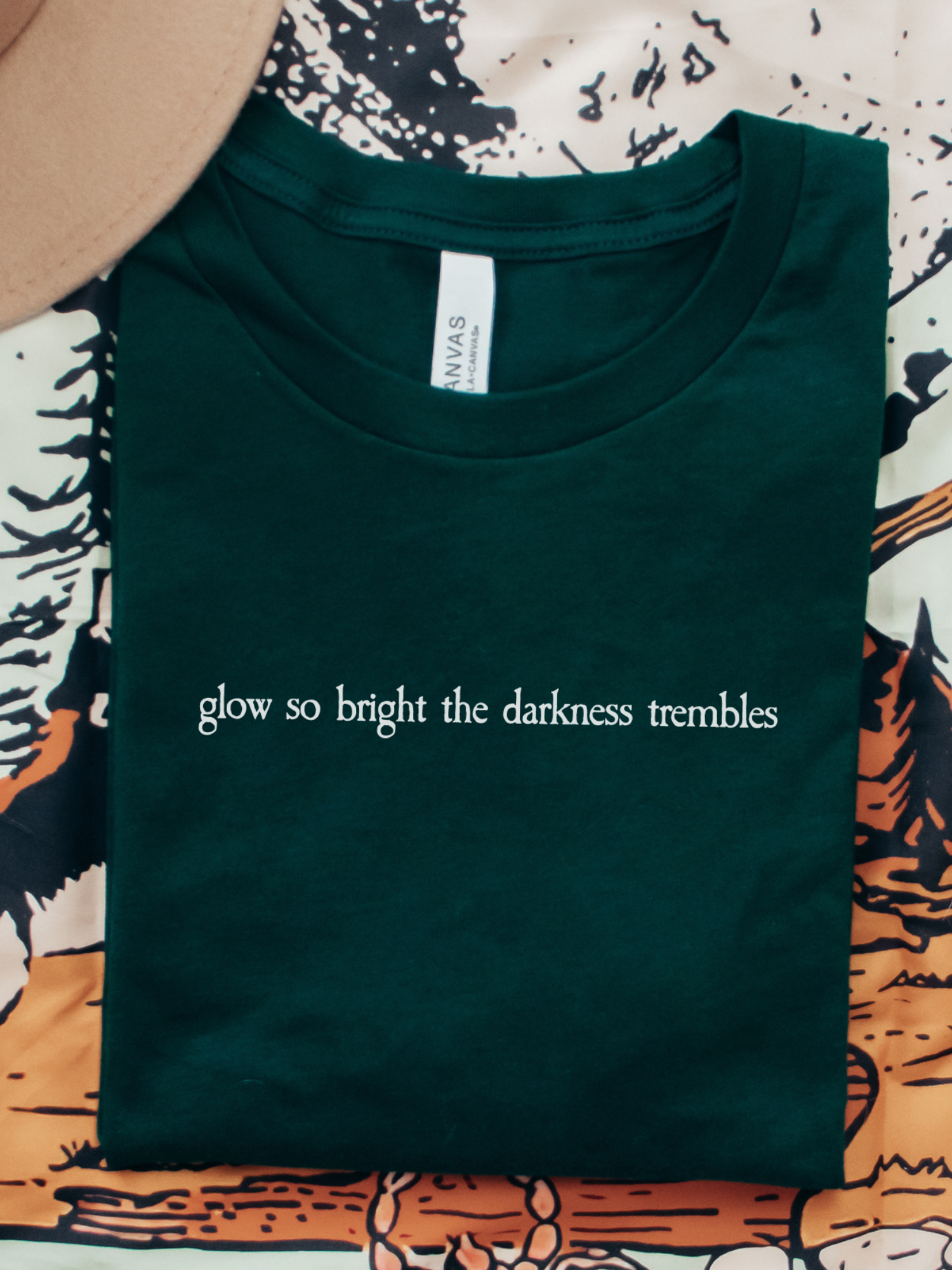 Forest green Bella Canvas shirt folded flat, printed with the quote “glow so bright the darkness trembles” in white fantasy serif font, placed on a fantasy-style illustrated background with a beige hat