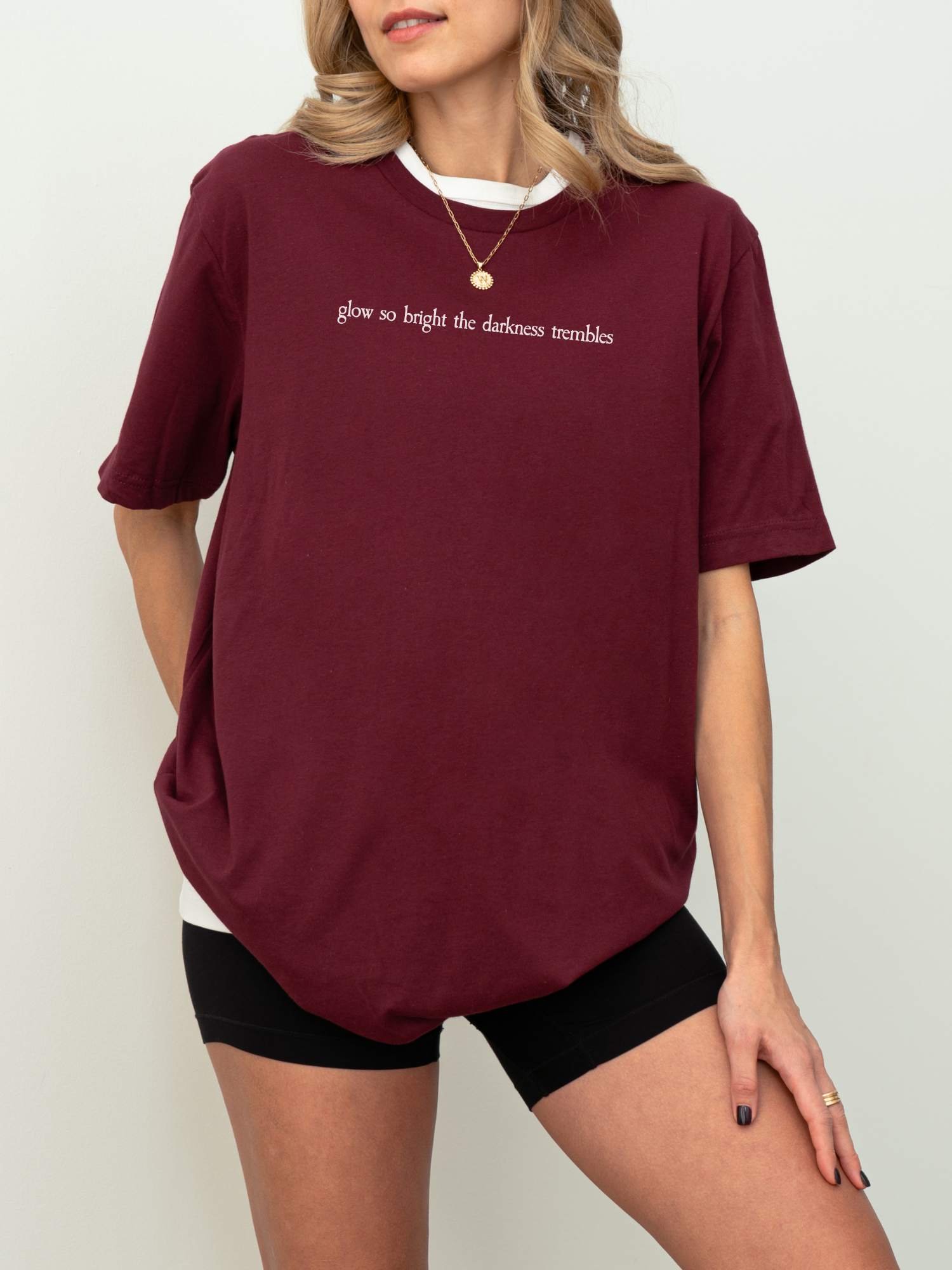 Maroon Bella Canvas shirt with the quote “glow so bright the darkness trembles” in white serif lettering, styled on a model wearing biker shorts and gold jewelry